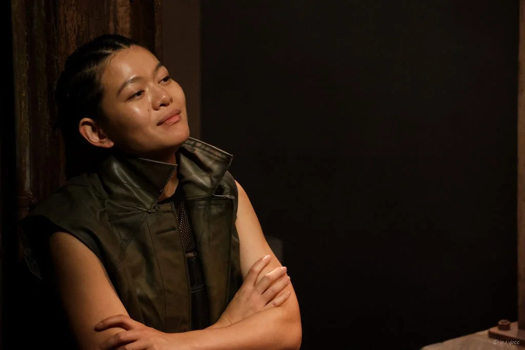 A young woman with braided hair, wearing a sleeveless black leather vest, leaning against a wooden wall with arms crossed, smirking slightly in a dimly lit room.