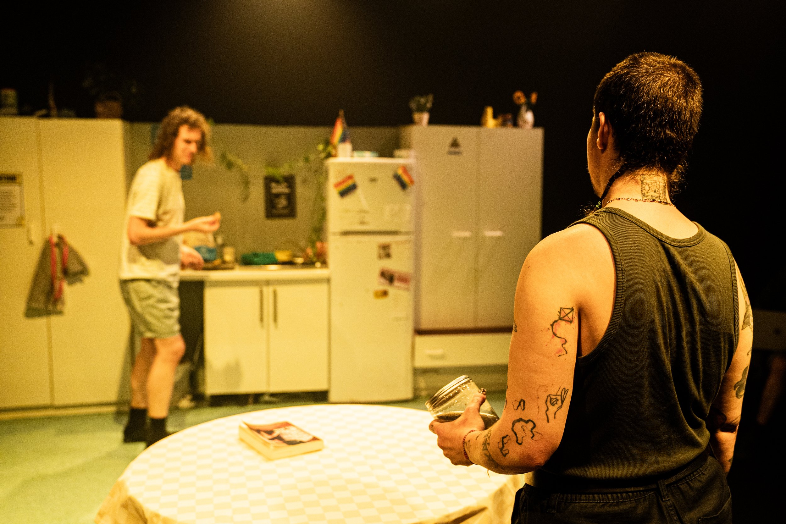 Two people in a kitchen with yellow cabinets, one with tattoos holding a jar, the other with curly hair preparing food, decorated with rainbow flags.