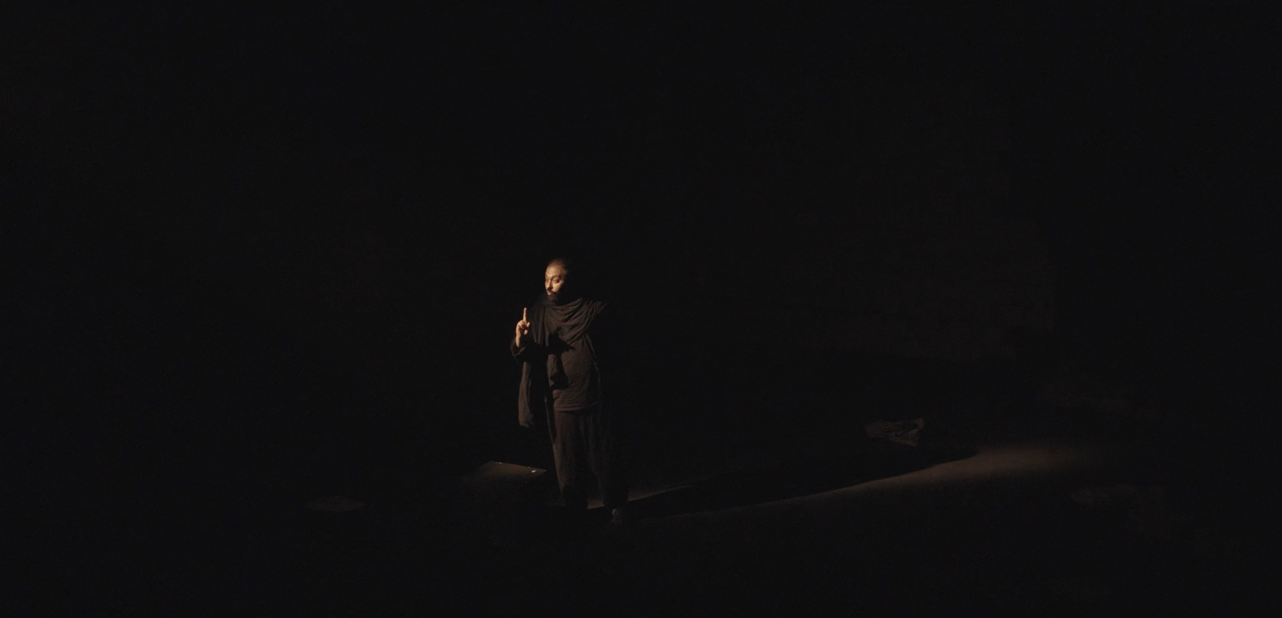 Person dressed in black standing in a spotlight in a dark room, making a peace sign with one hand.