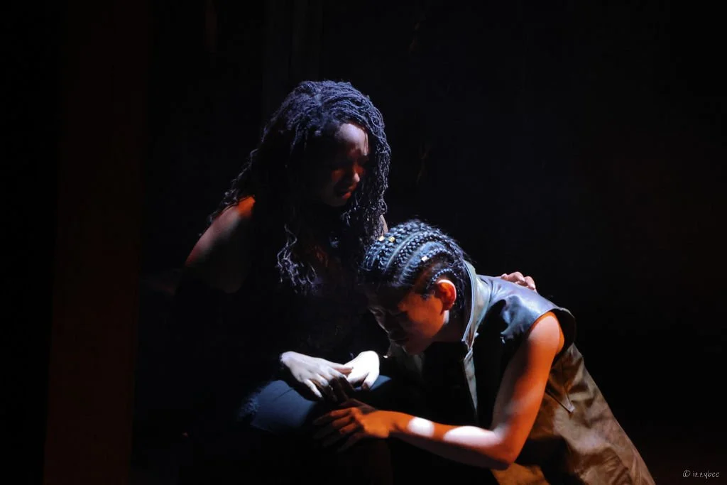 A woman with curly hair and a young boy with braided hair in a dark setting, possibly a theatrical performance, with the woman looking down at the boy who appears to be seeking comfort.