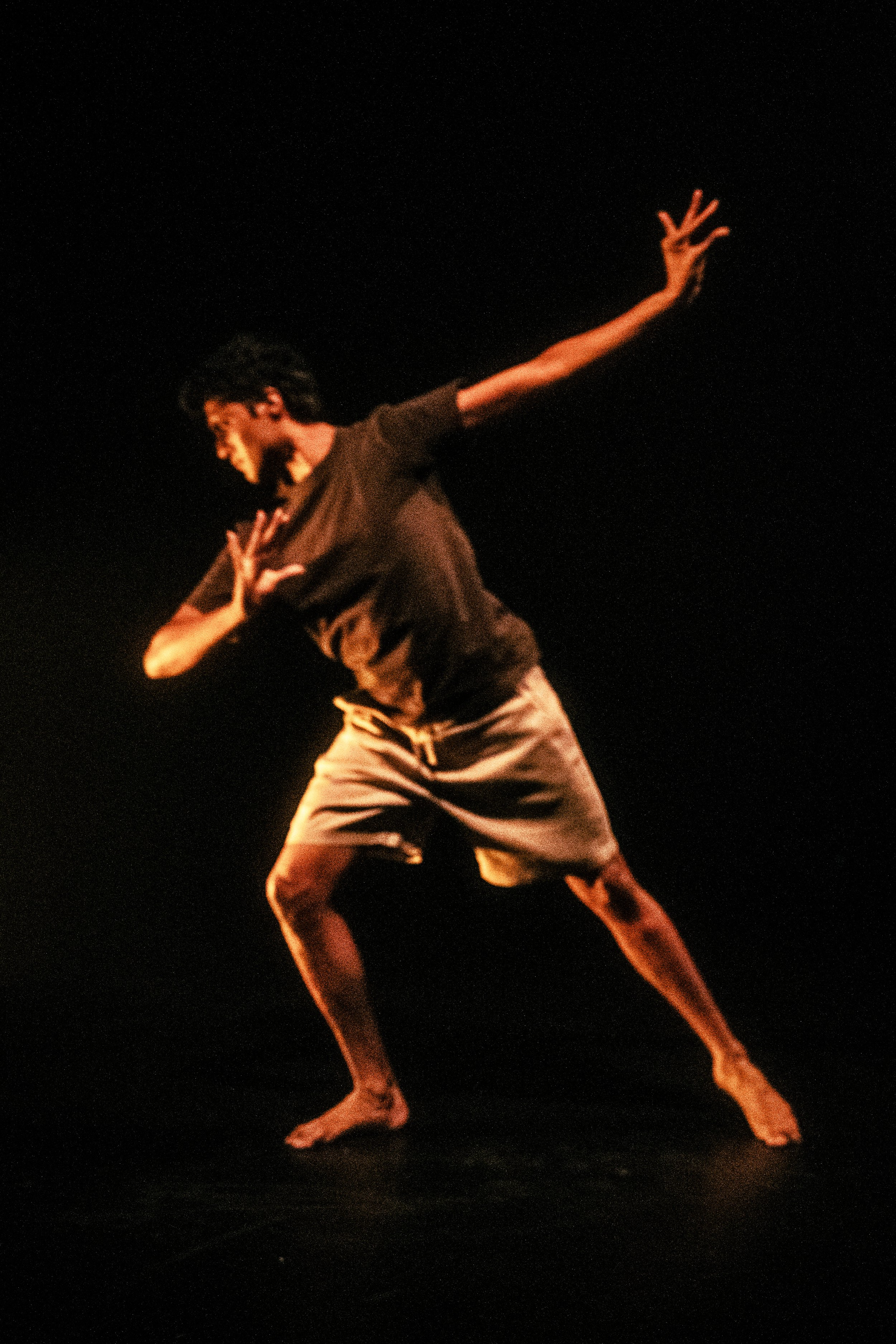 A person performing a dance move on a dark stage, with one arm extended behind and the other across its chest, wearing a brown t-shirt and tan shorts.