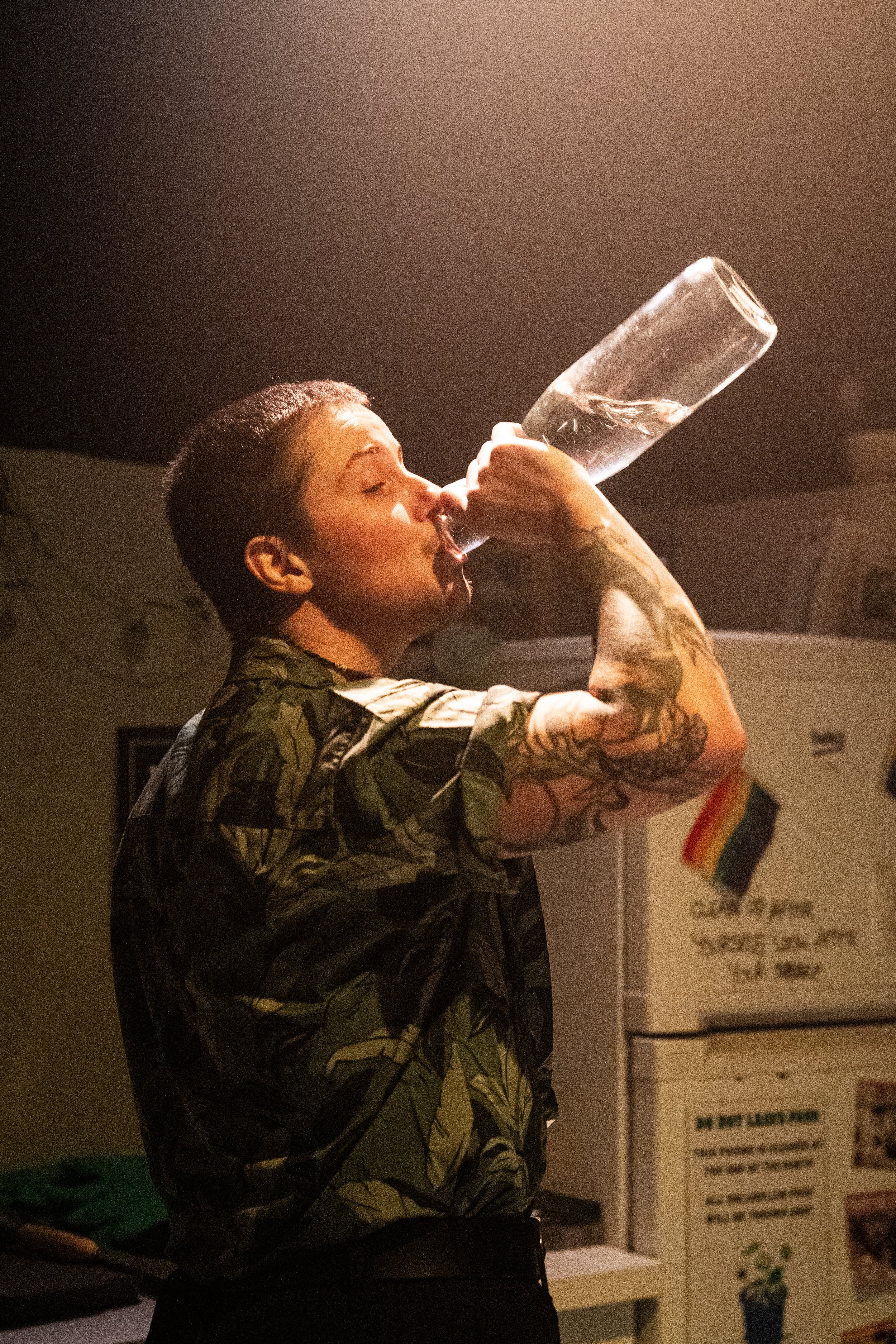 A person with short hair and tattoos on their arm is wearing a patterned shirt and drinking from a clear bottle in a room with a refrigerator decorated with a rainbow pride flag magnet.