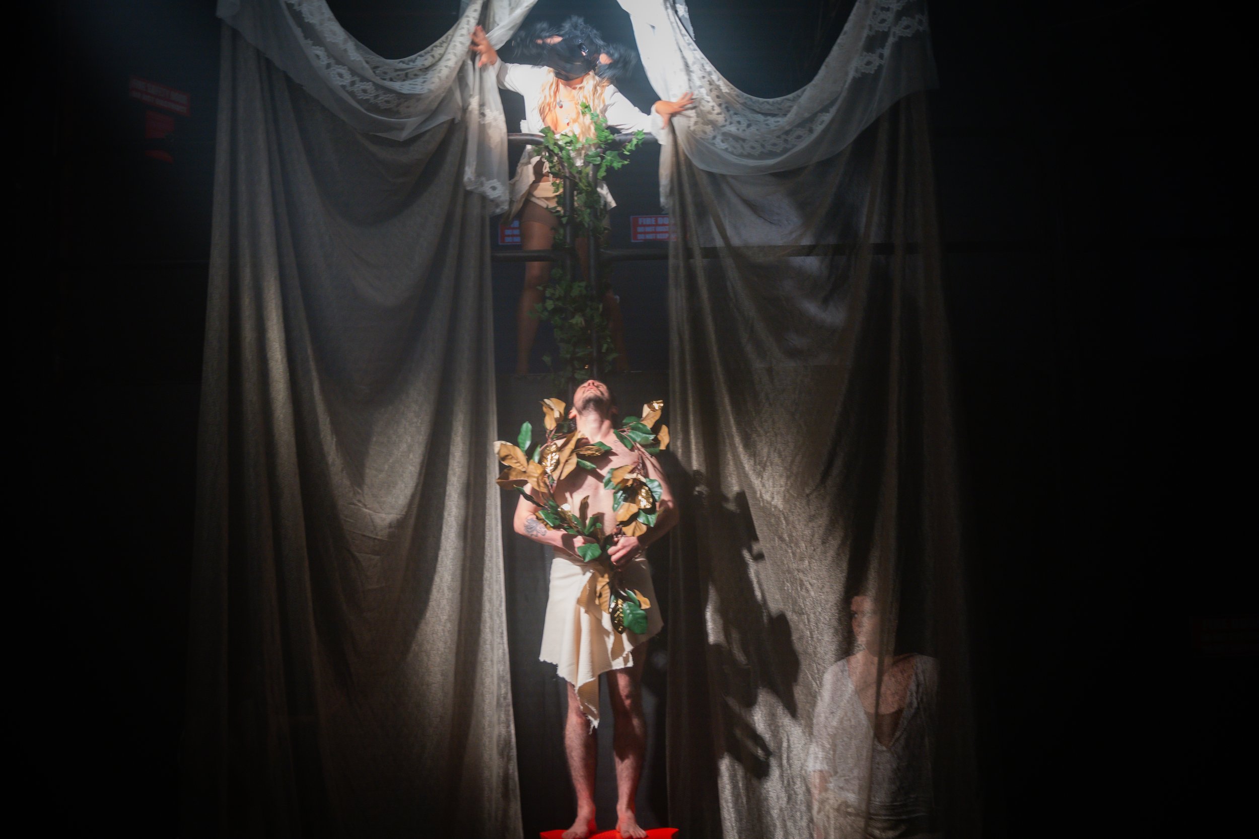 Stage performance with two actors, one with leafy costume, one at window with lace curtains and plants.