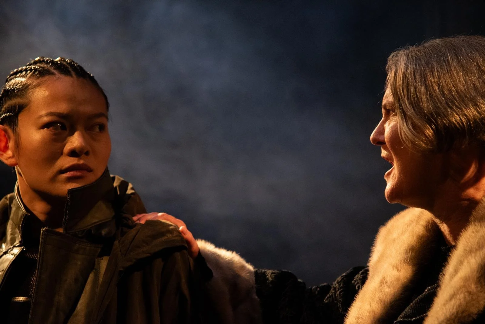 Two people engaged in a serious conversation; a woman with short hair wearing a fur garment and an older woman with braided hair in a dark environment.