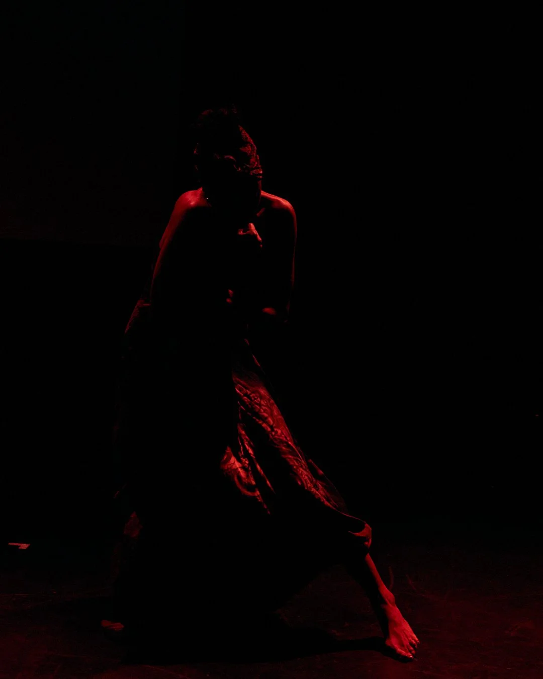A person illuminated by red light with a dark background, appearing to be in a dramatic pose.