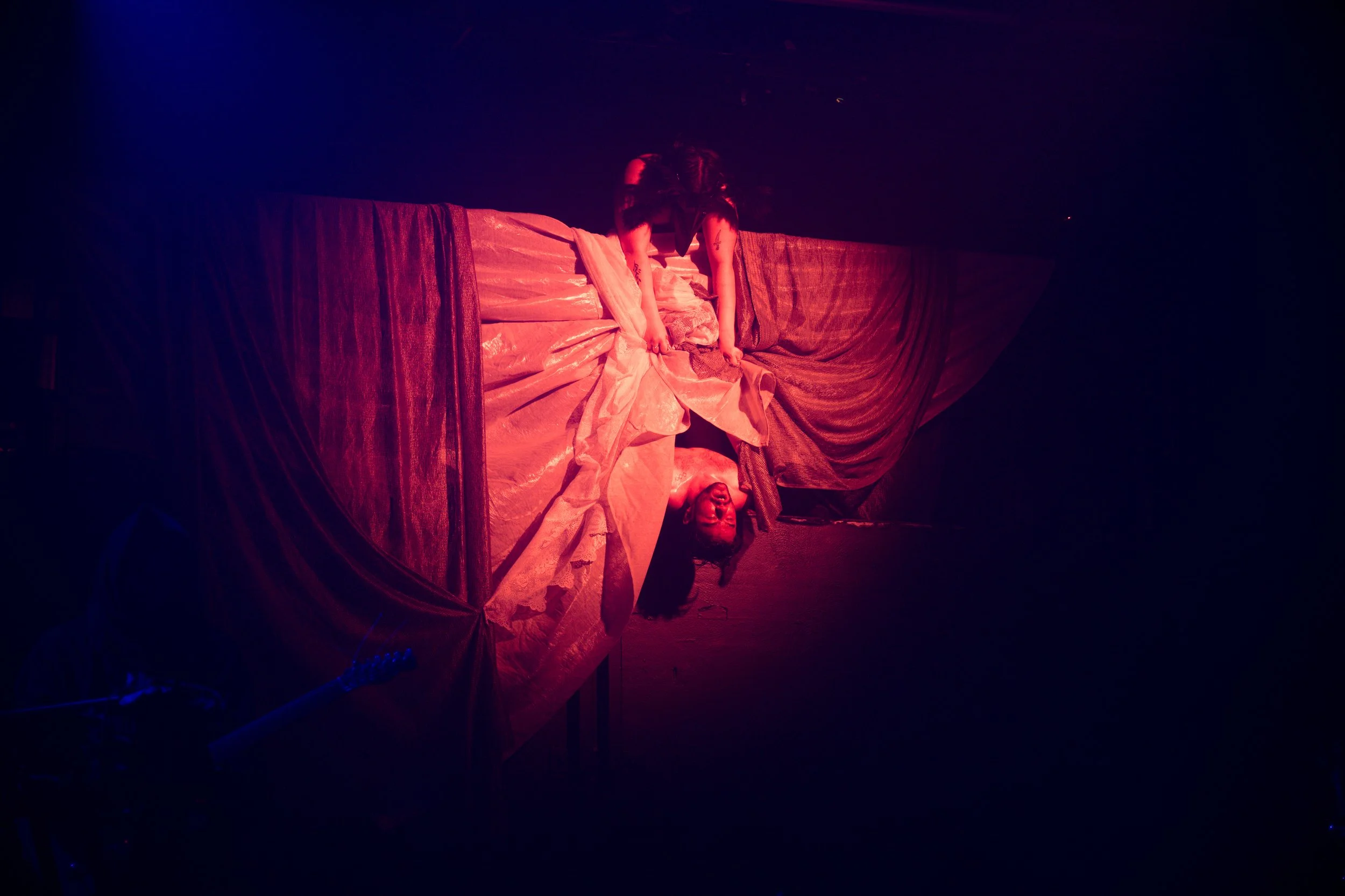 Two performers on a stage show a dramatic act with draped fabric; the person on top is reaching down while the other is looking up with an expressive face, all under dim, colored lighting.