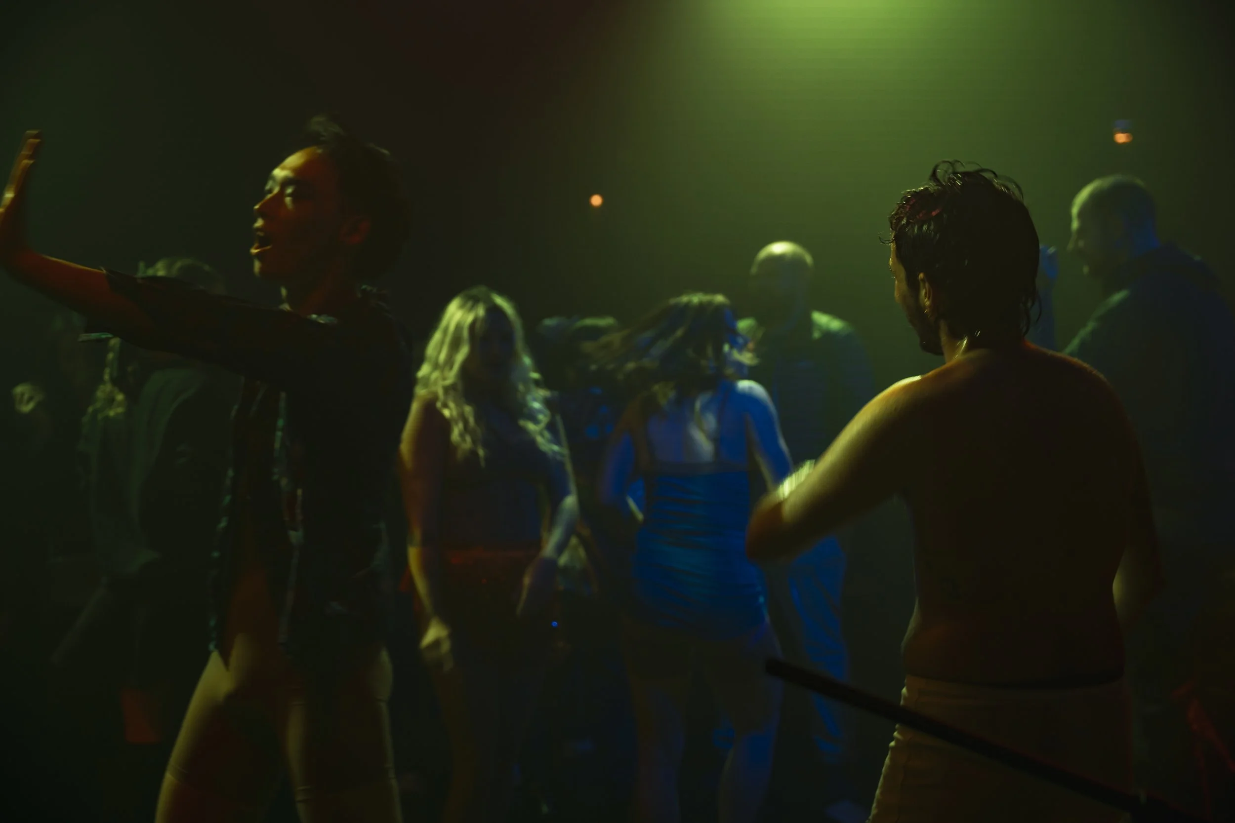 People dancing in a dark, colorful nightclub with green, yellow, and blue lighting.