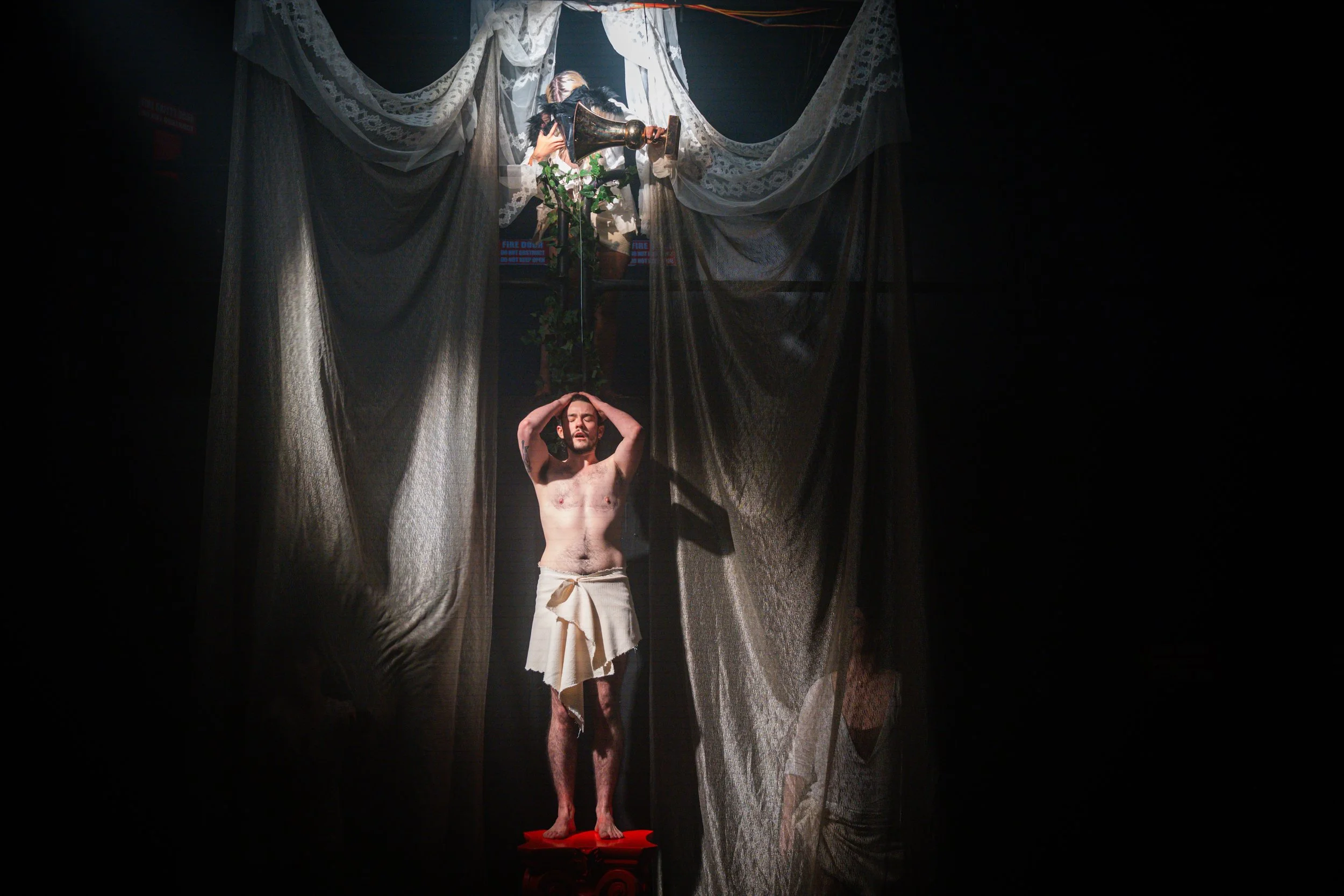 A man standing on a red surface with a cloth wrapped around his waist, holding his head with both hands, in front of dark curtains. Above him, on a platform, another person appears to be interacting with a vintage-style gramophone amid potted plants,