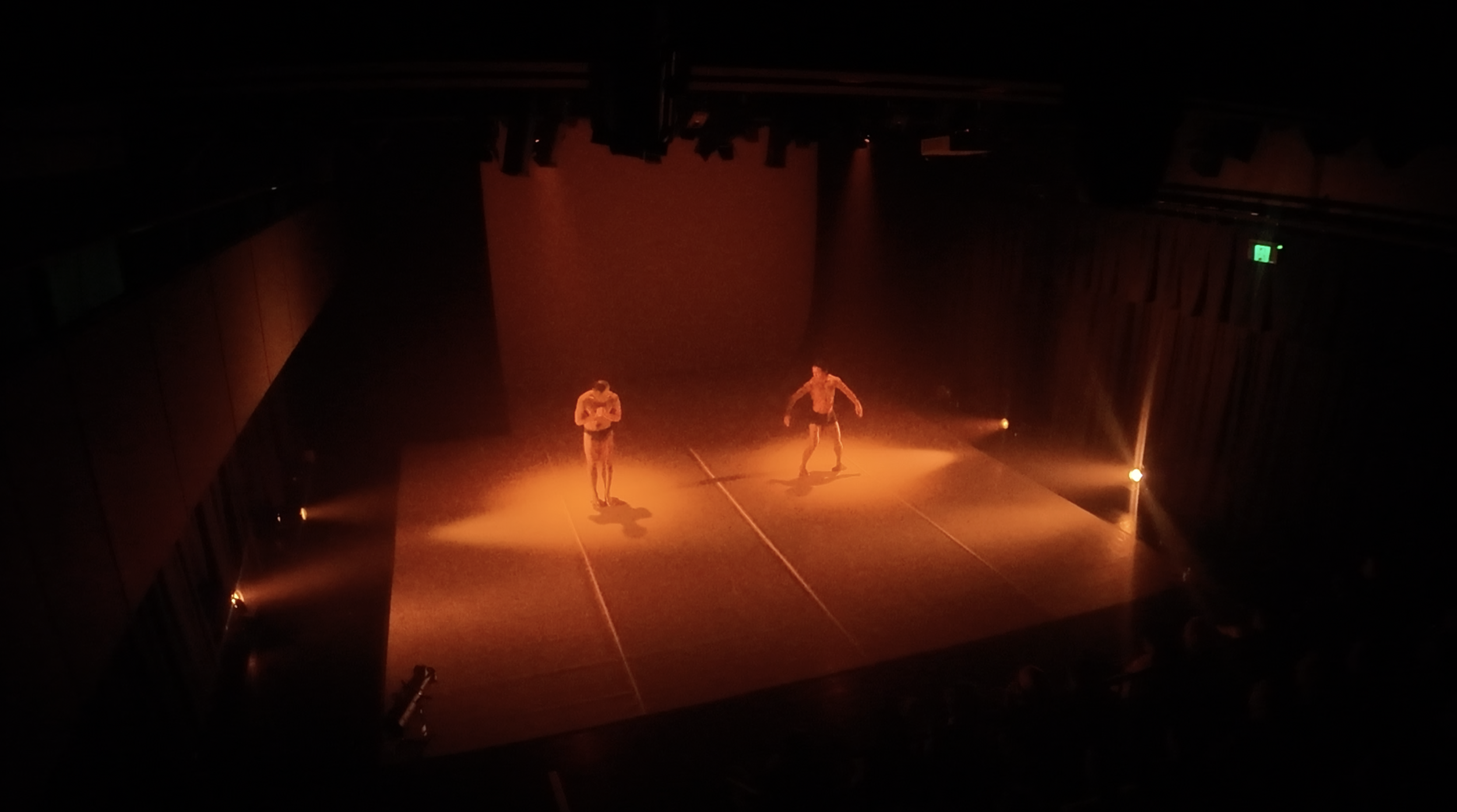 Two performers on a dimly lit stage with orange lighting, one reading a book and the other dancing, in a theater setting.
