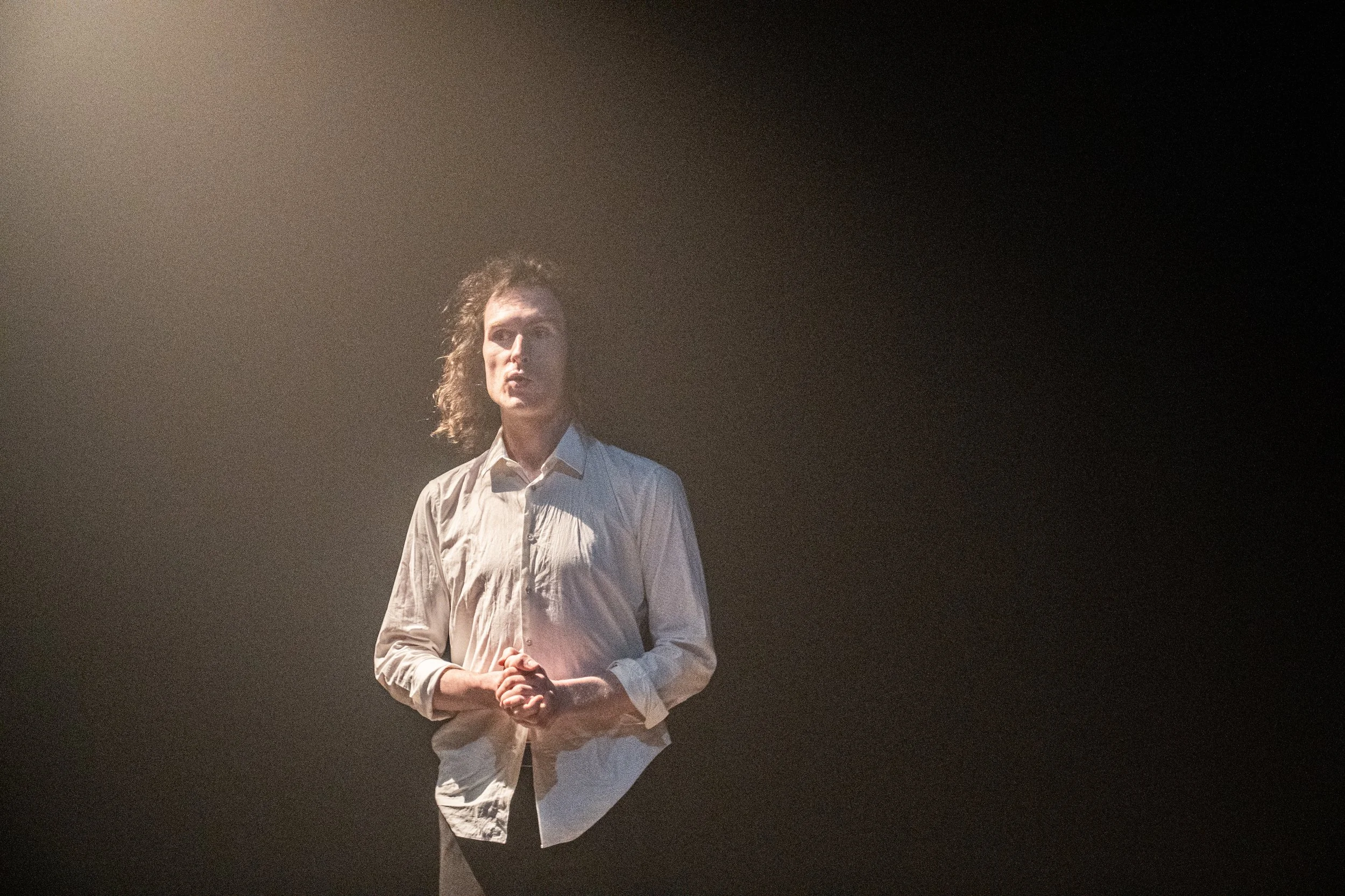 A woman with curly hair in a white shirt stands on a dark stage with a spotlight shining on her.