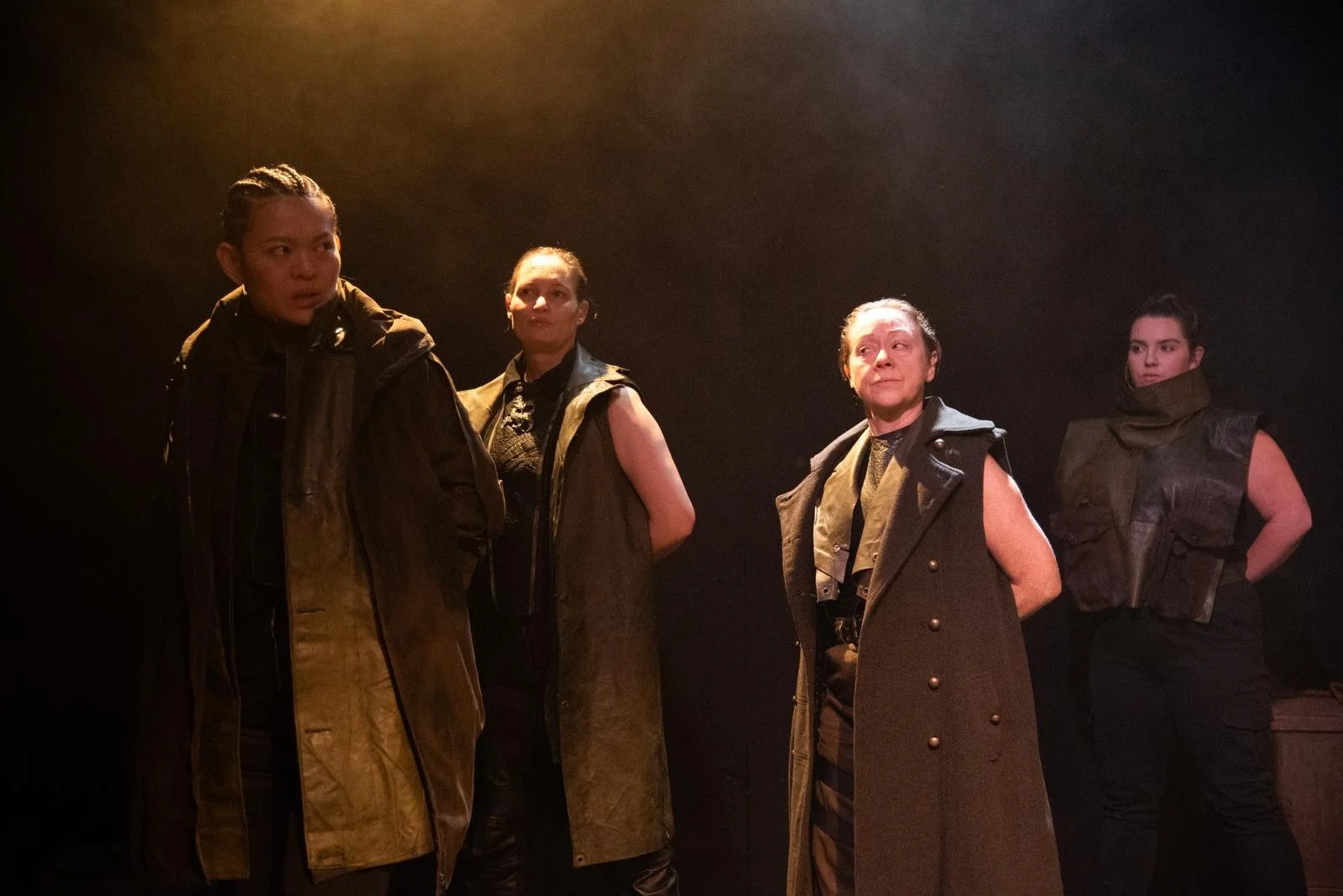 Four women standing on a dark stage, dressed in armored and military-style clothing, with serious expressions.