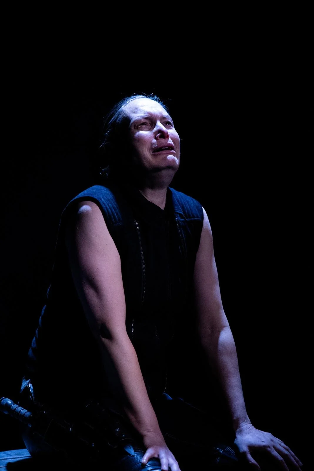 A woman with short hair crying and looking upward, illuminated by a spotlight against a dark background.