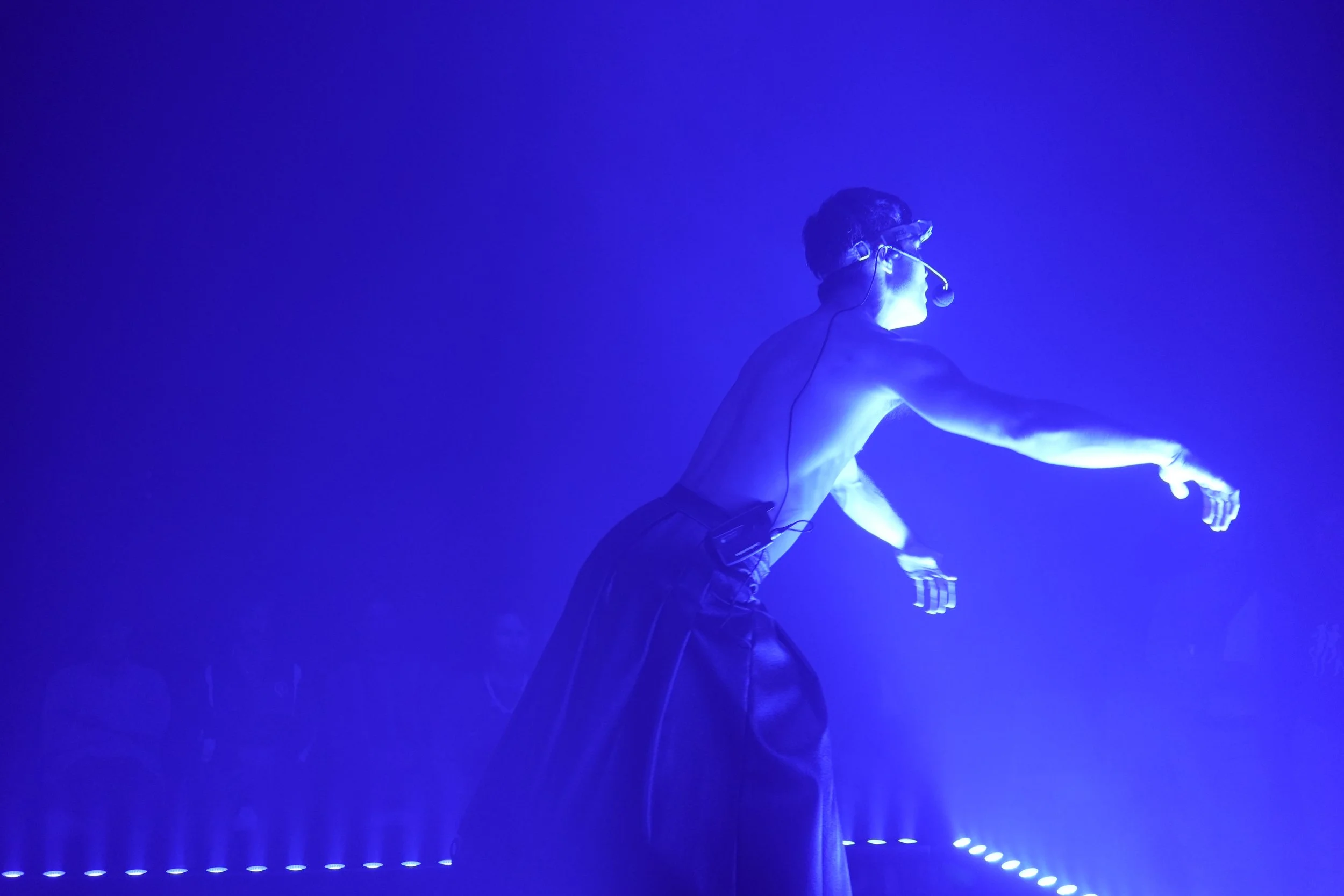 A shirtless man wearing a headset and a kilt performing a dance in blue lighting.