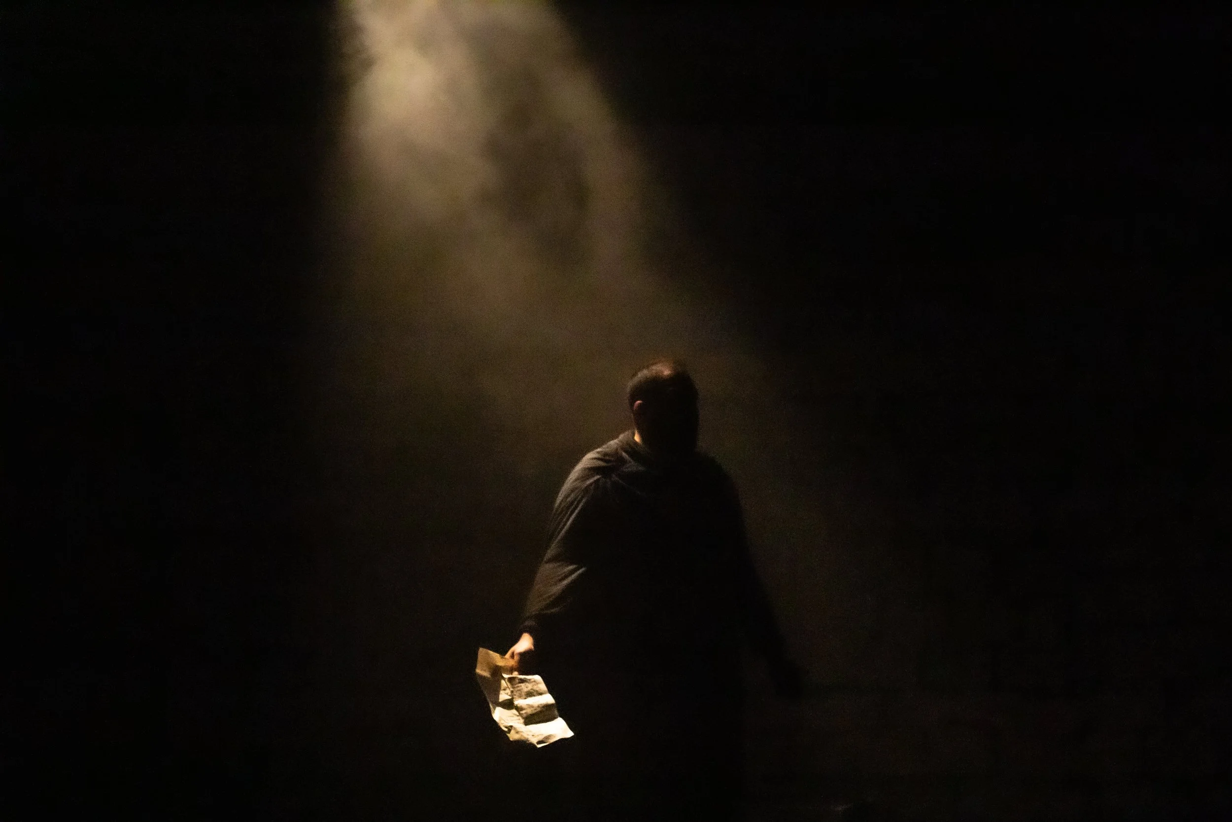 Person standing in a dark room with a spotlight shining down, holding an object in one hand.