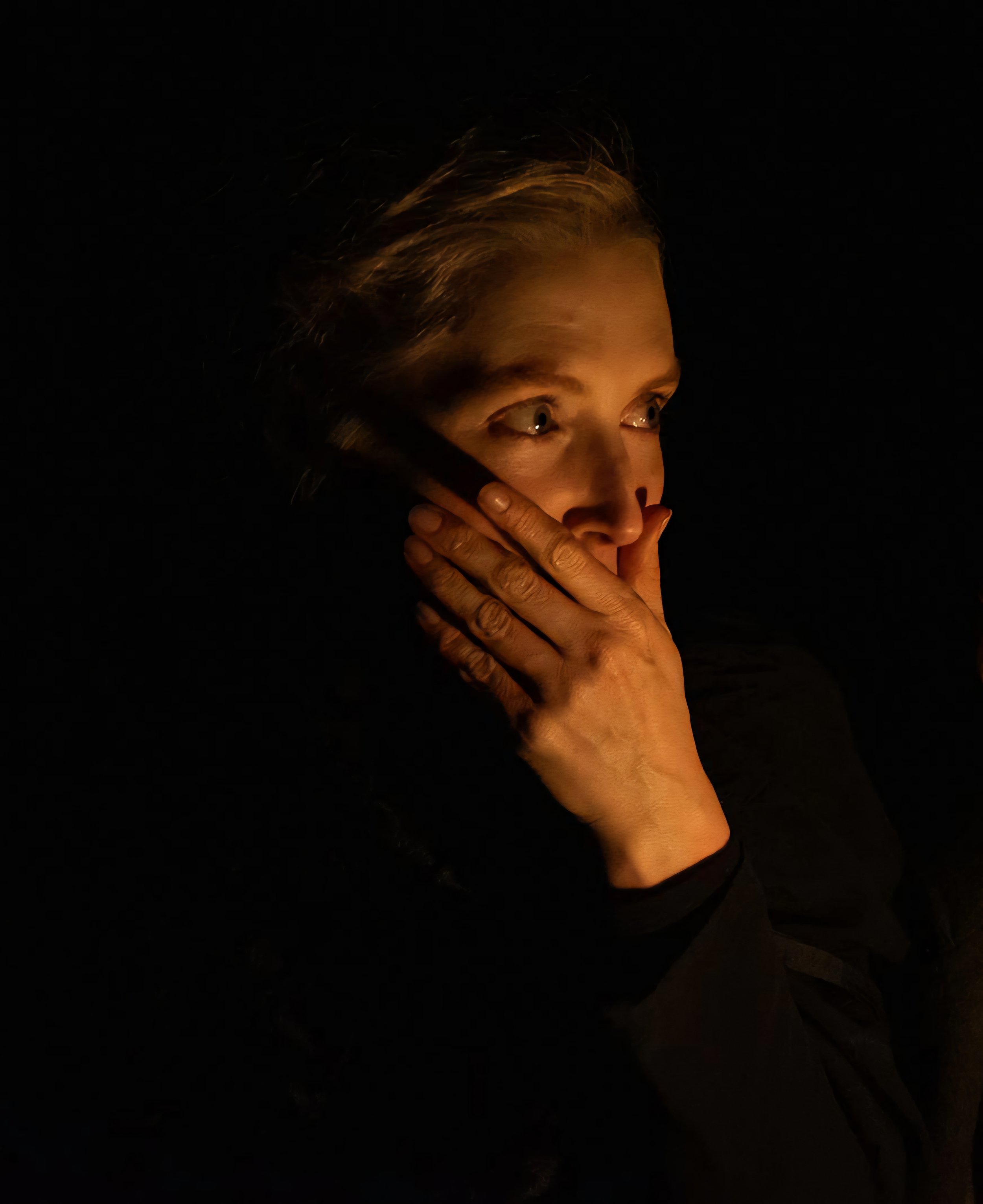 A woman with blonde hair in a dark setting, illuminated by warm light, covering her mouth with her right hand and looking to the side.