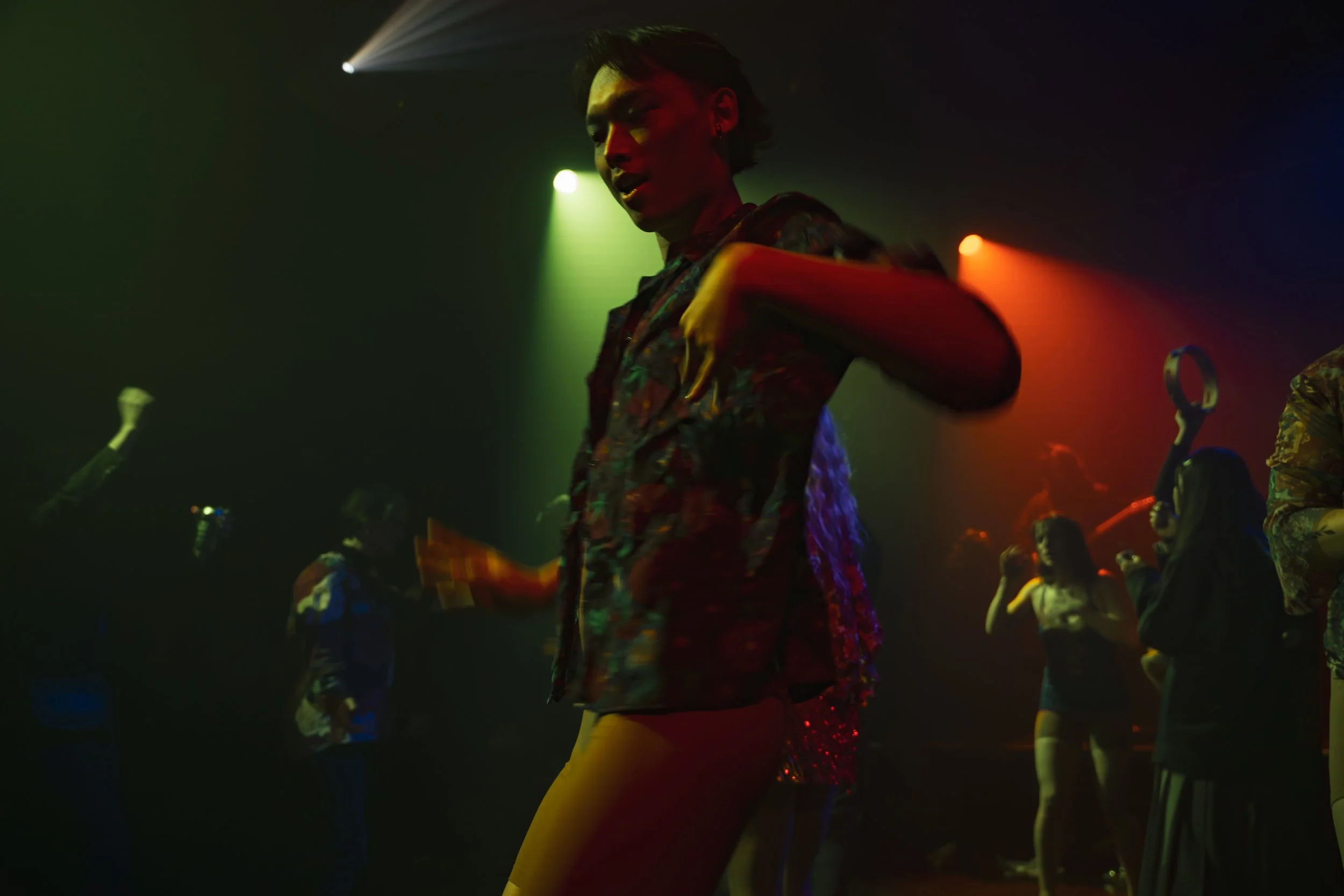People dancing in a dark club with colorful lighting, including a woman in the foreground with short hair and a patterned shirt.