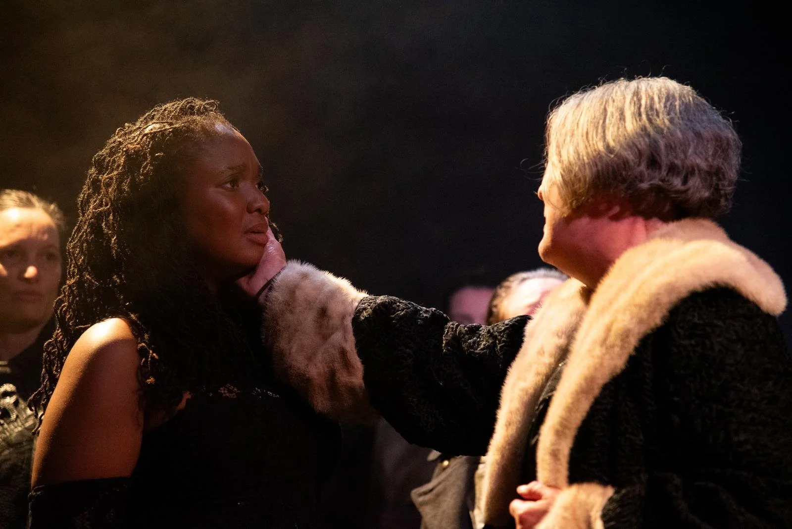 A woman with curly hair and dark clothing holds the face of an older woman with short hair and a black coat with cream-colored fur at the collar, appearing emotional in a dimly lit setting.