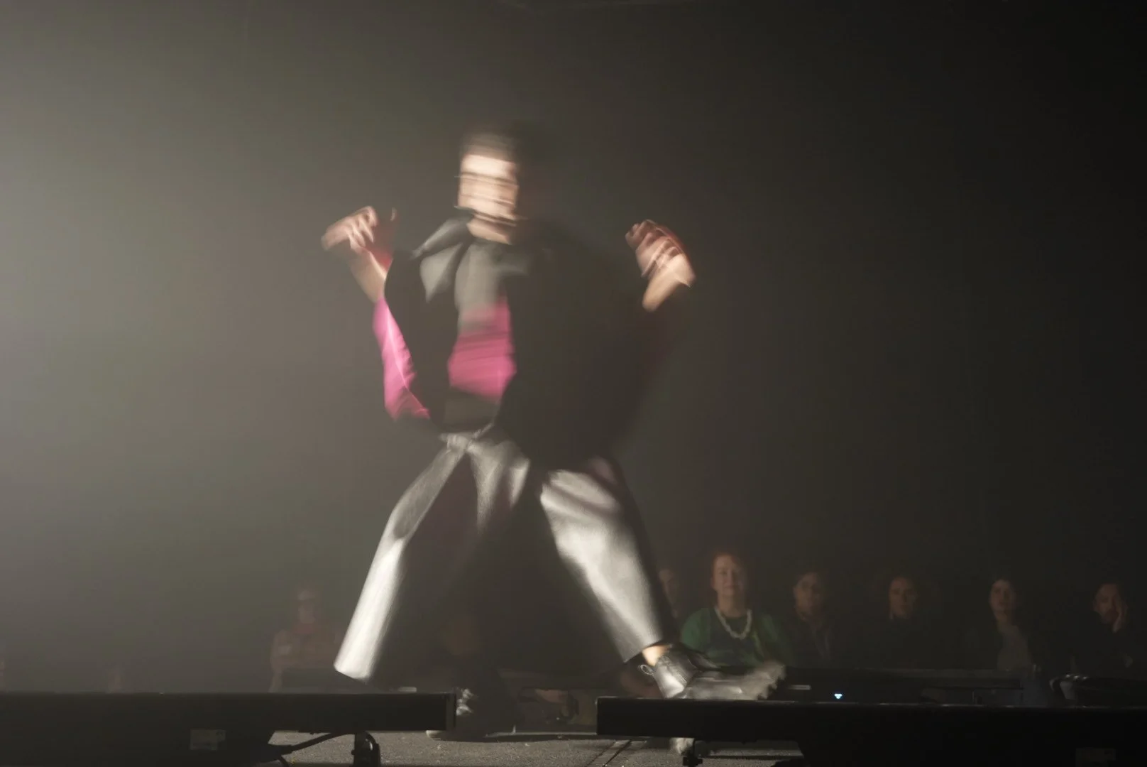Person dancing on stage with audience in background, stage lights casting a glow.