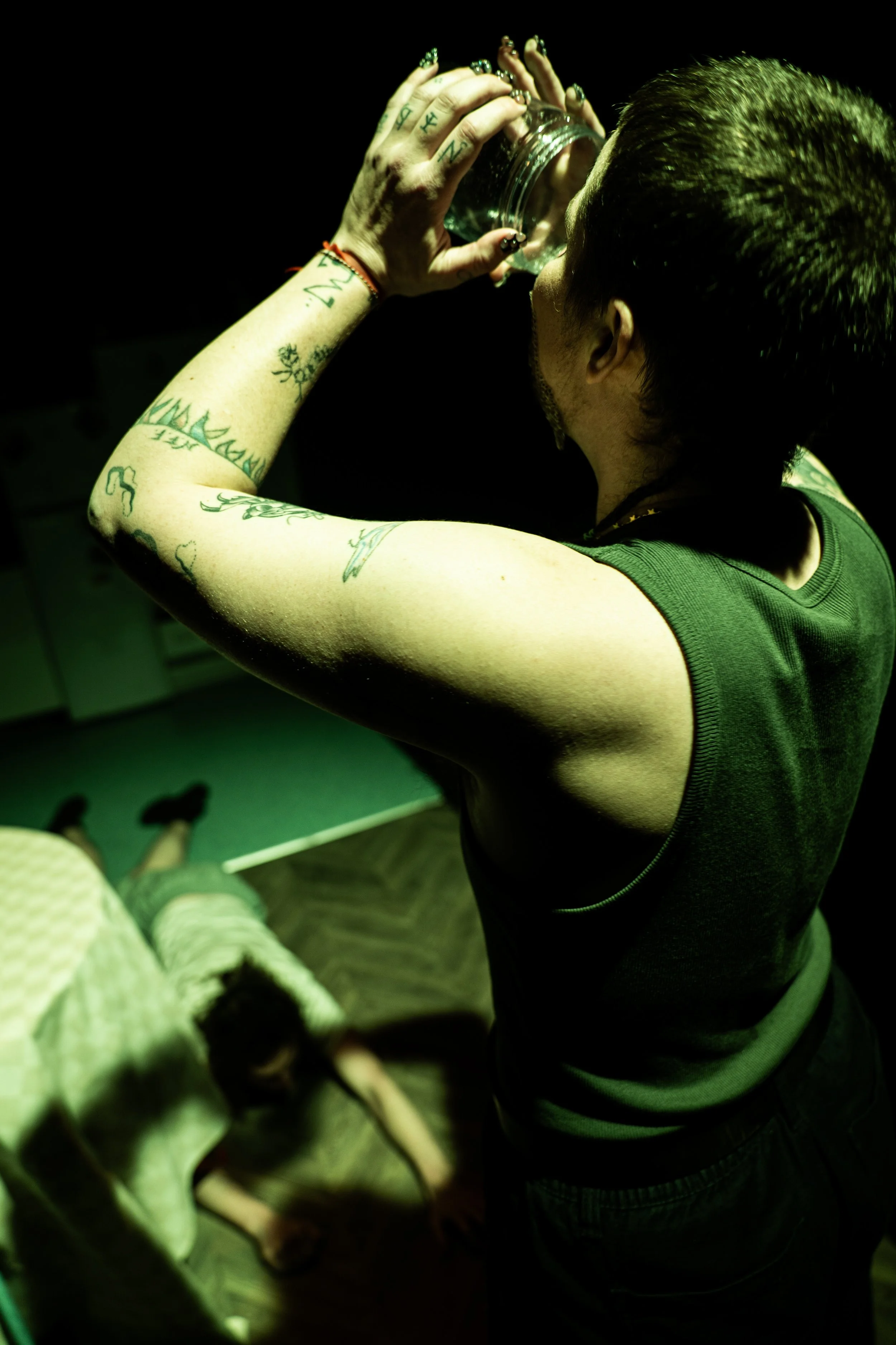 A person with tattoos drinking from a glass, seen from behind, wearing a sleeveless dark shirt in a dimly lit room, with a dog on a chair in the background.