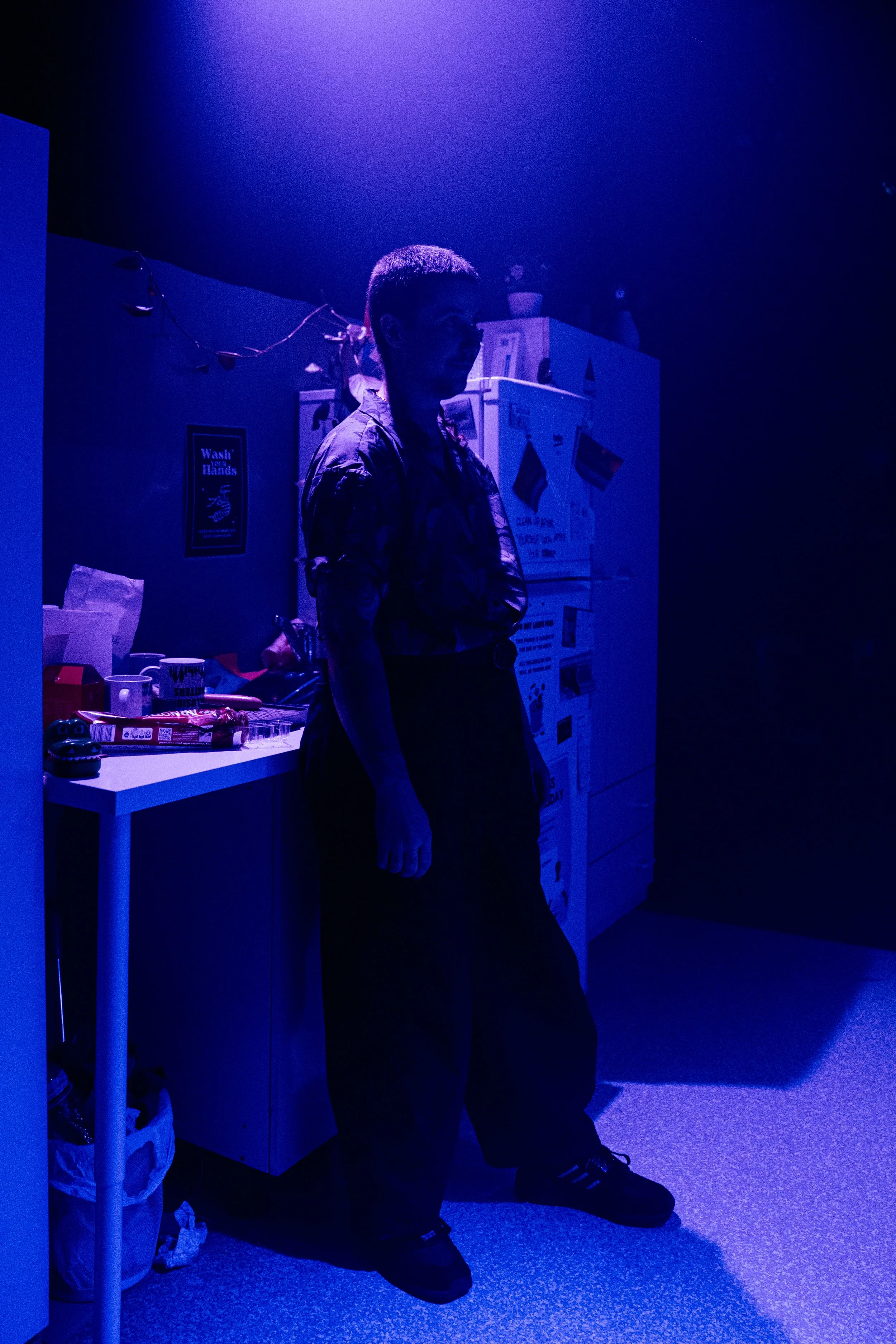 A person standing in a dimly lit room illuminated by purple light, with a refrigerator and a table with various objects behind them.