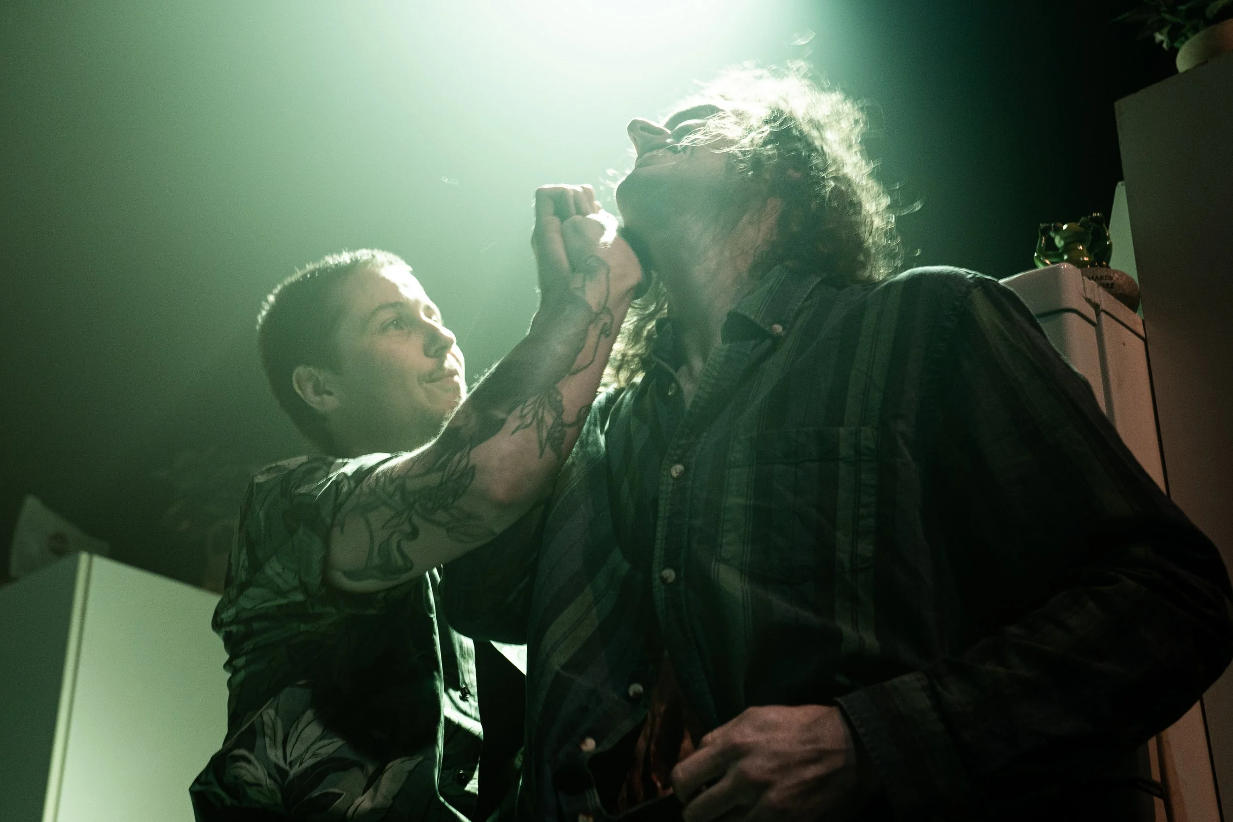 Tattoed young person with short hair, holding a microphone, performing in front of a man with long hair and a plaid shirt in a dimly lit room.
