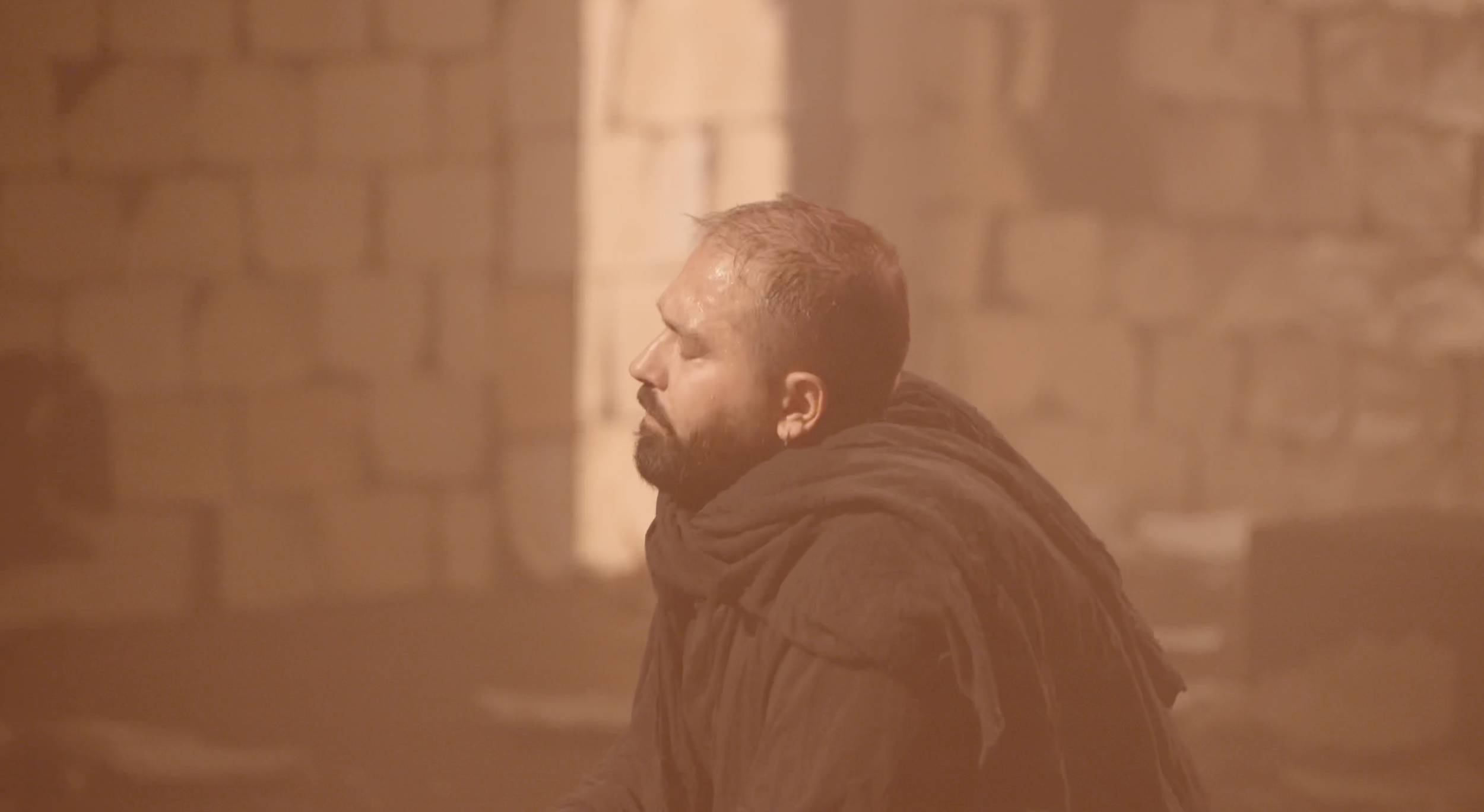 A man with short hair and a beard, dressed in a dark hoodie, sitting with his eyes closed in what appears to be a smoke-filled or foggy environment with warm lighting and brick walls in the background.