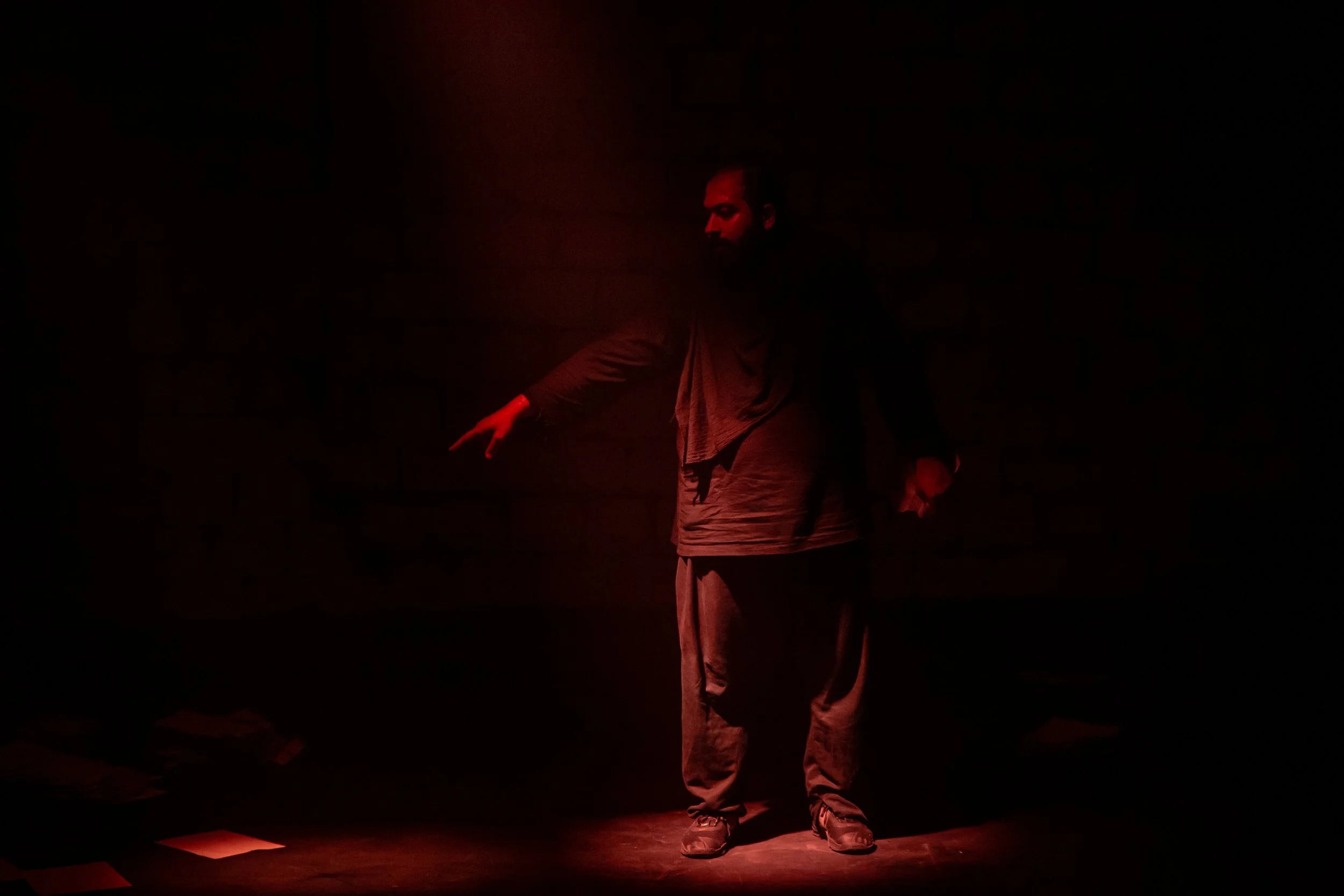 A person standing on a dark stage illuminated by a faint red light, with their head slightly bowed and one arm extended downwards.