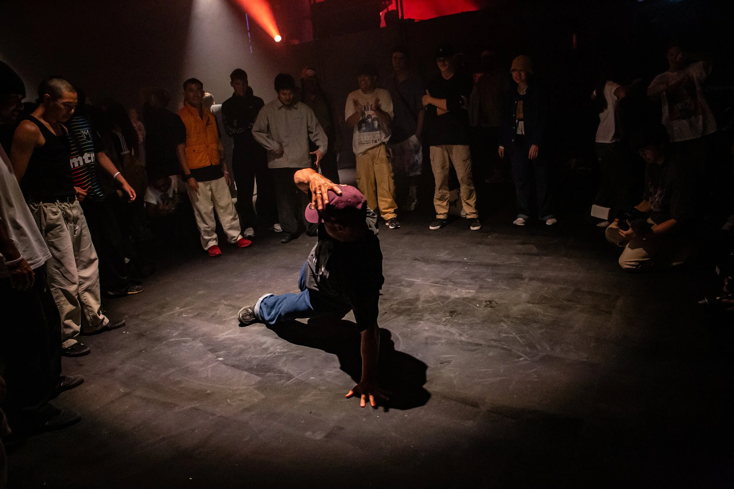 A person breakdancing on the floor in front of an audience in a dimly lit room with red and orange lights.