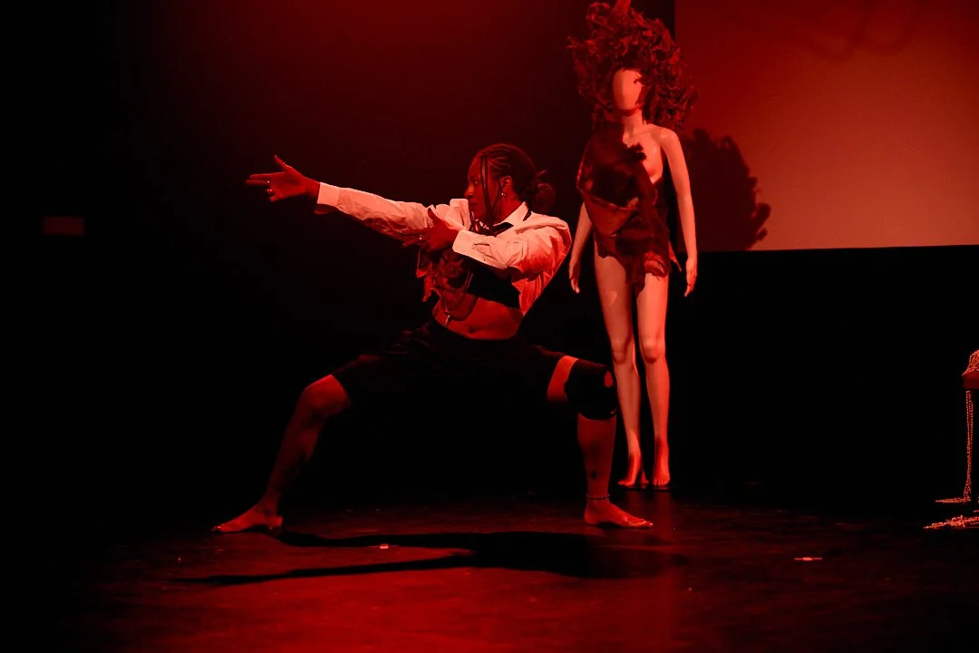 A person performing a dance move on stage with a red lighting backdrop, wearing black shorts with red straps, a white shirt, and a black vest, with hands extended to the side. A mannequin with long curly hair and a dress is in the background.