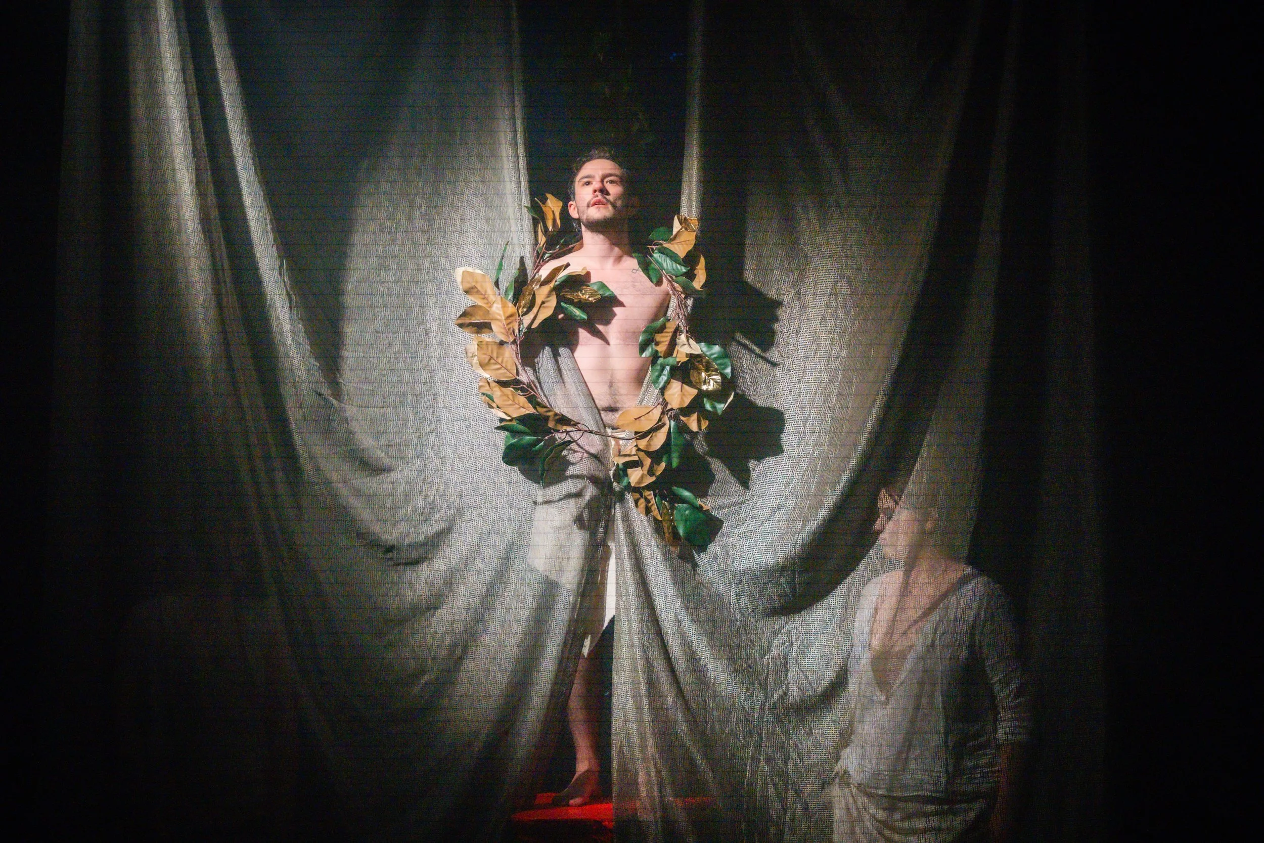 A man standing on a stage, topless, wearing a leafy wreath around his neck and shoulders, with gray trousers, partially obscured by dark curtains. A woman is sitting on the floor, partly hidden behind the curtain, observing him.