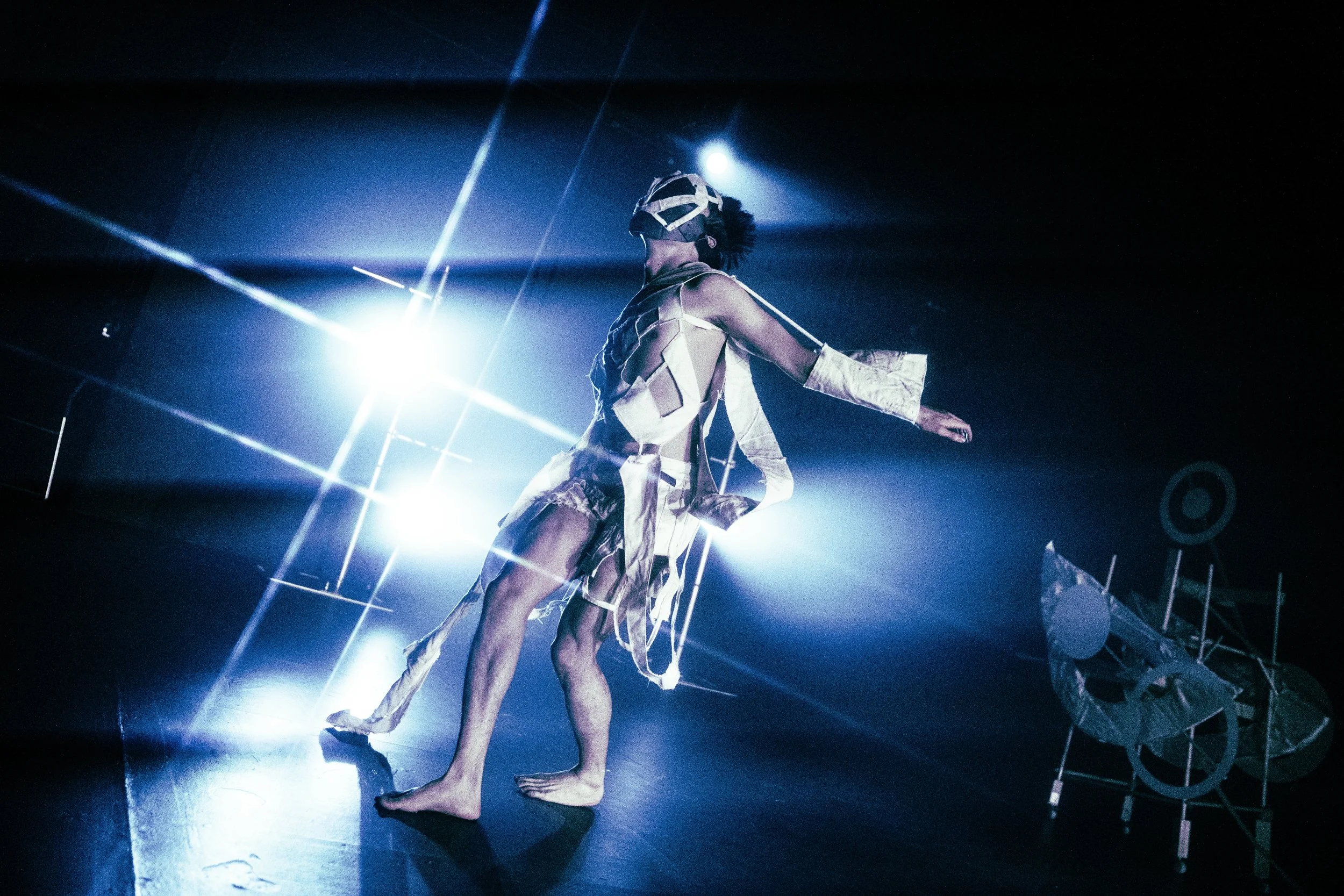 Performer dressed in a beige and white costume with torn fabric, barefoot, on stage with blue lighting and star-shaped light effects, with a white mechanical bicycle prop in the background.