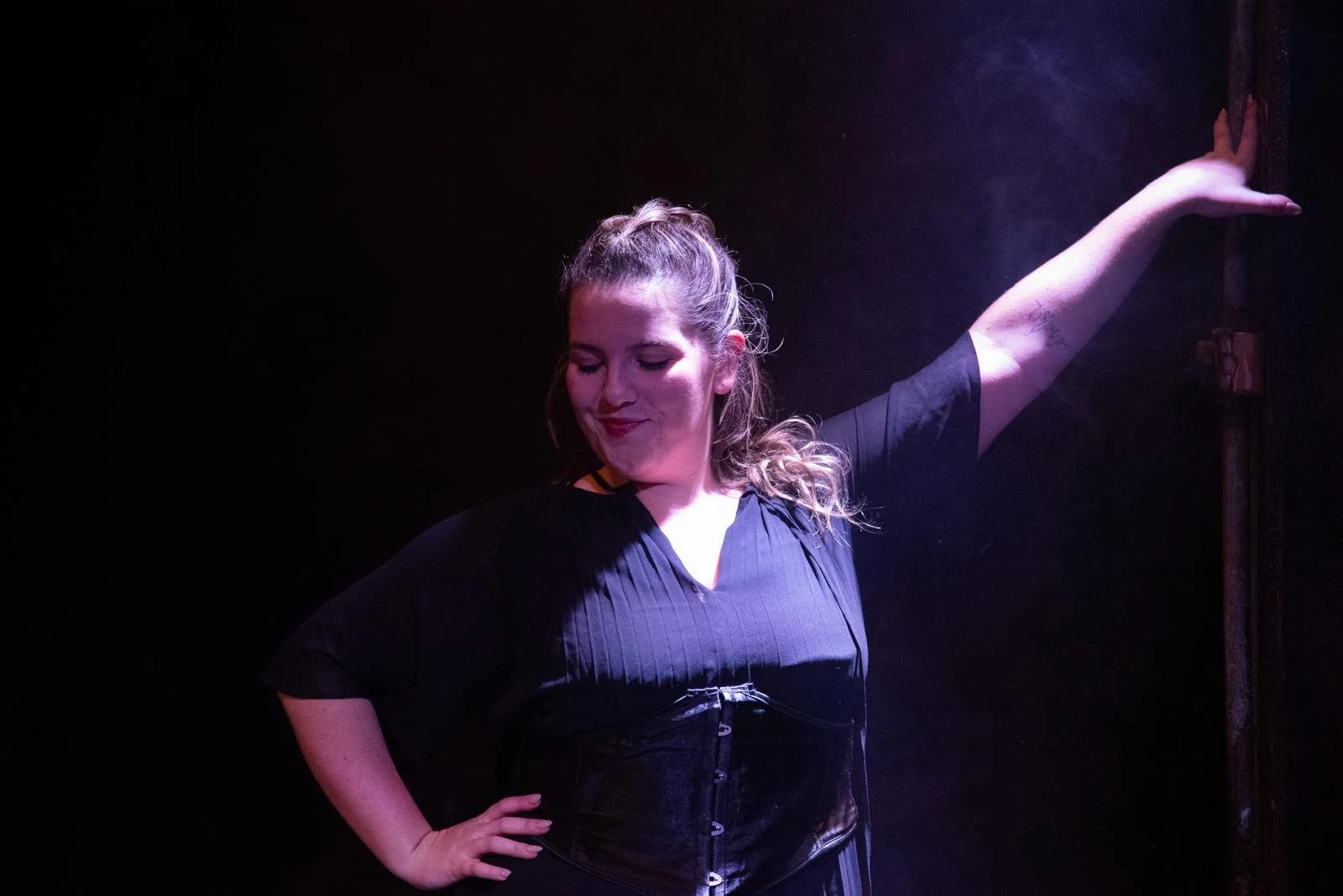 A woman with light skin and curly, light brown hair styled in a ponytail is striking a pose with one arm extended upward and the other on her hip, standing against a dark background illuminated by purple lighting.