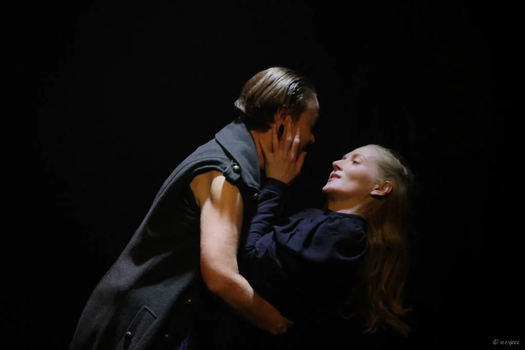 A man and woman in an intimate dance or embrace against a black background, looking into each other's eyes.