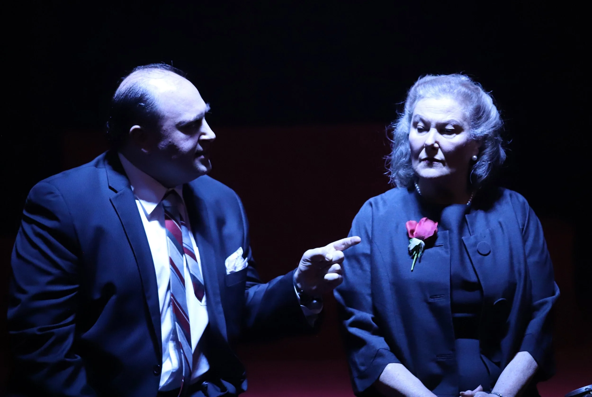 A man in a suit and tie is pointing his finger at an older woman with gray hair, who is wearing a dark jacket with a pink flower pin, in a dimly lit setting.