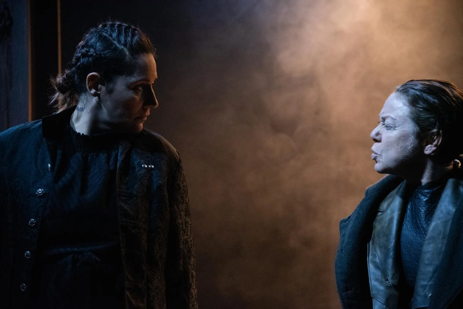 Two women face each other in a dark, smoky setting with contemplative expressions.