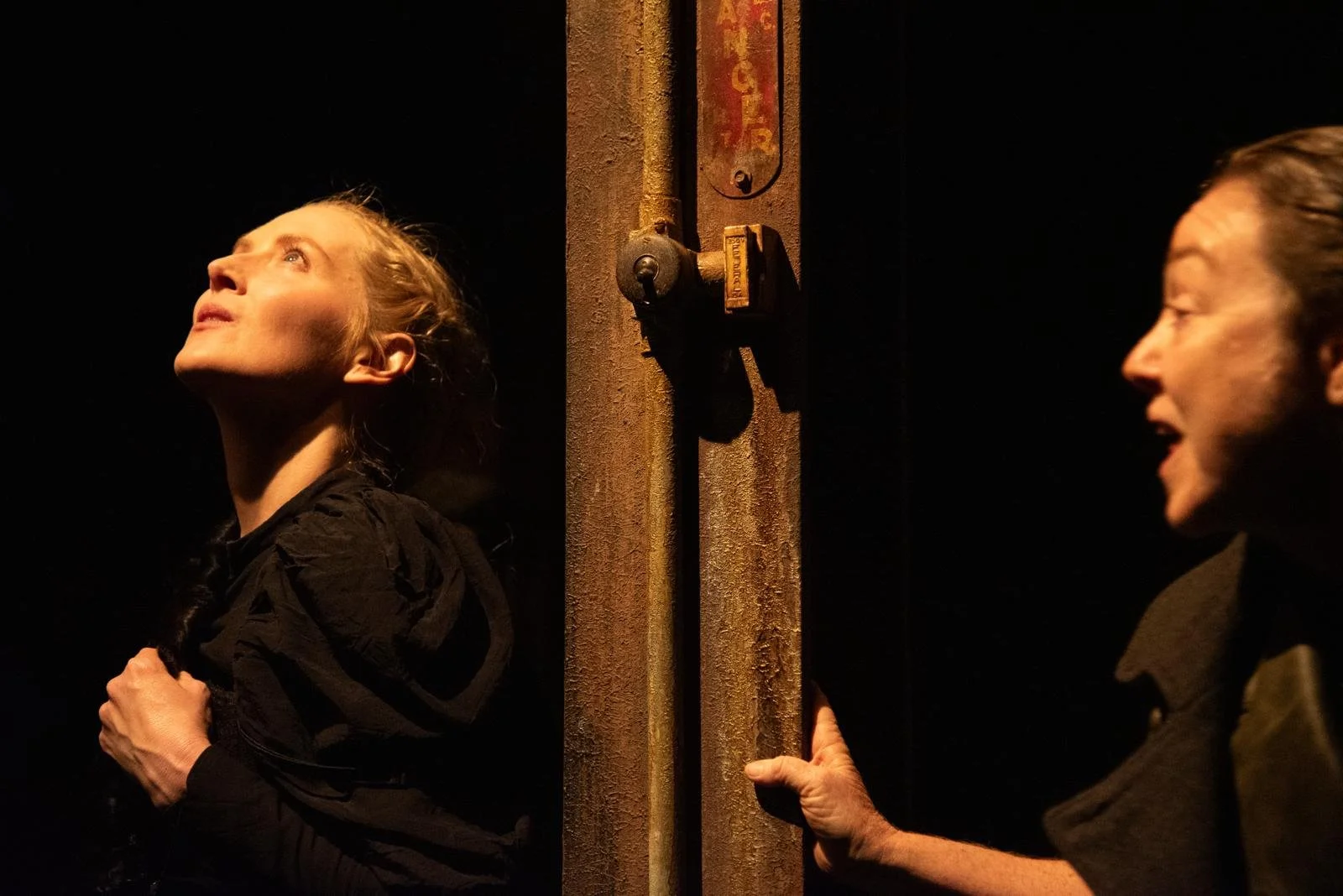 Two women on either side of a rusty metal door, one looking upward with a concerned expression, the other holding the door handle.