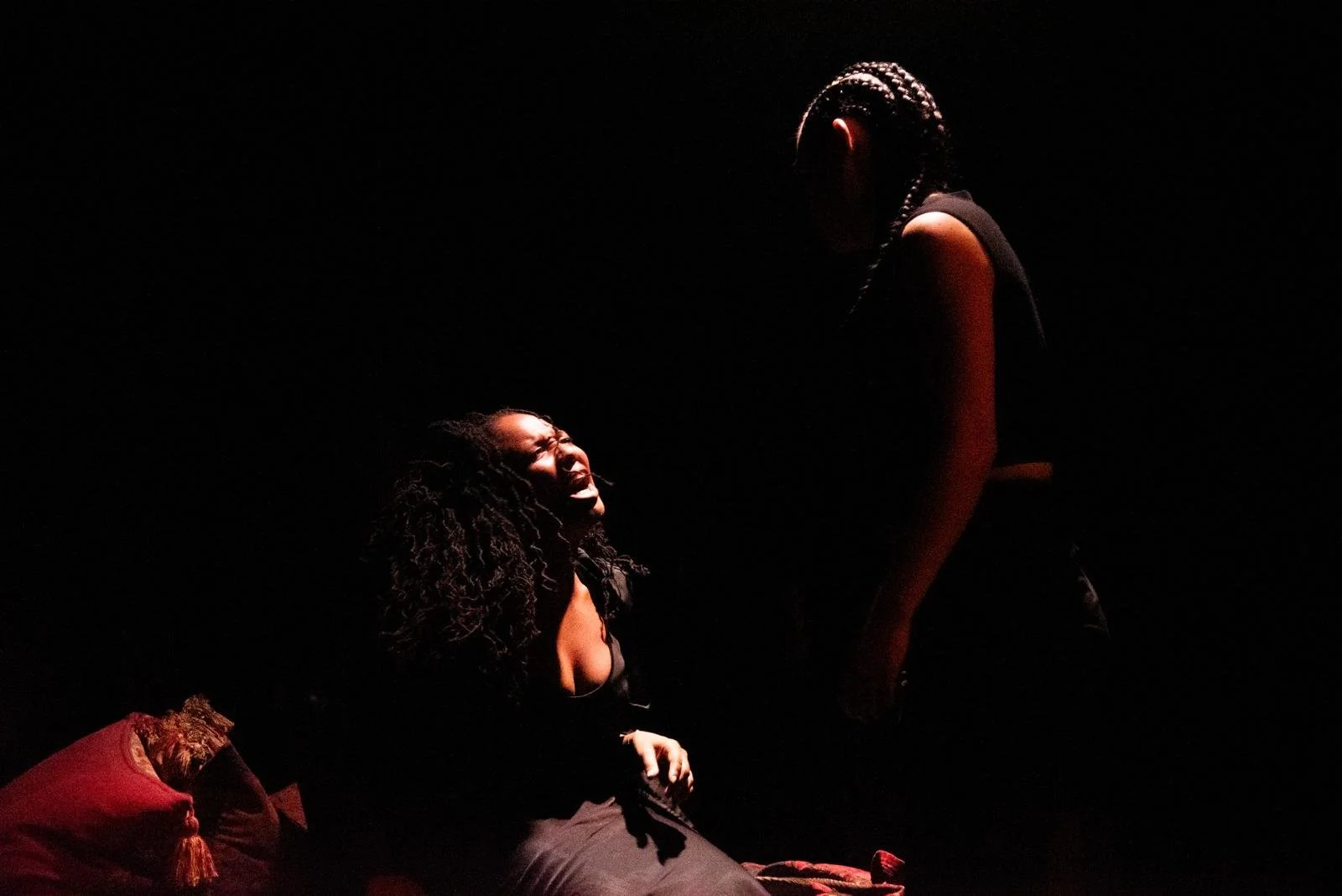 A woman kneeling with her eyes closed and mouth open, appearing to be singing or crying, illuminated by a soft light against a dark background. Another woman, standing and facing her, is partially lit and has braided hair.