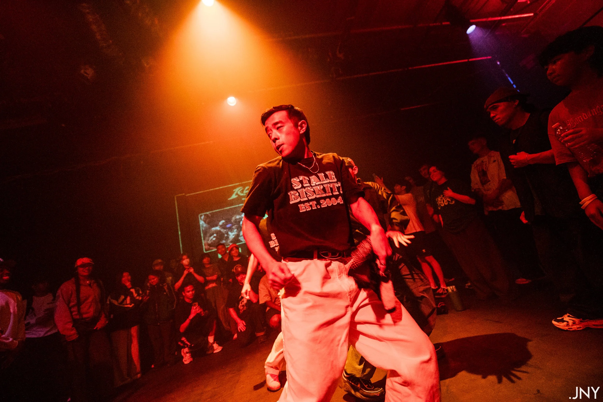 A young man dancing at a party or club with a group of people gathered around him in a dimly lit space with red and orange lighting. The background shows a screen with a logo and people watching the dance.