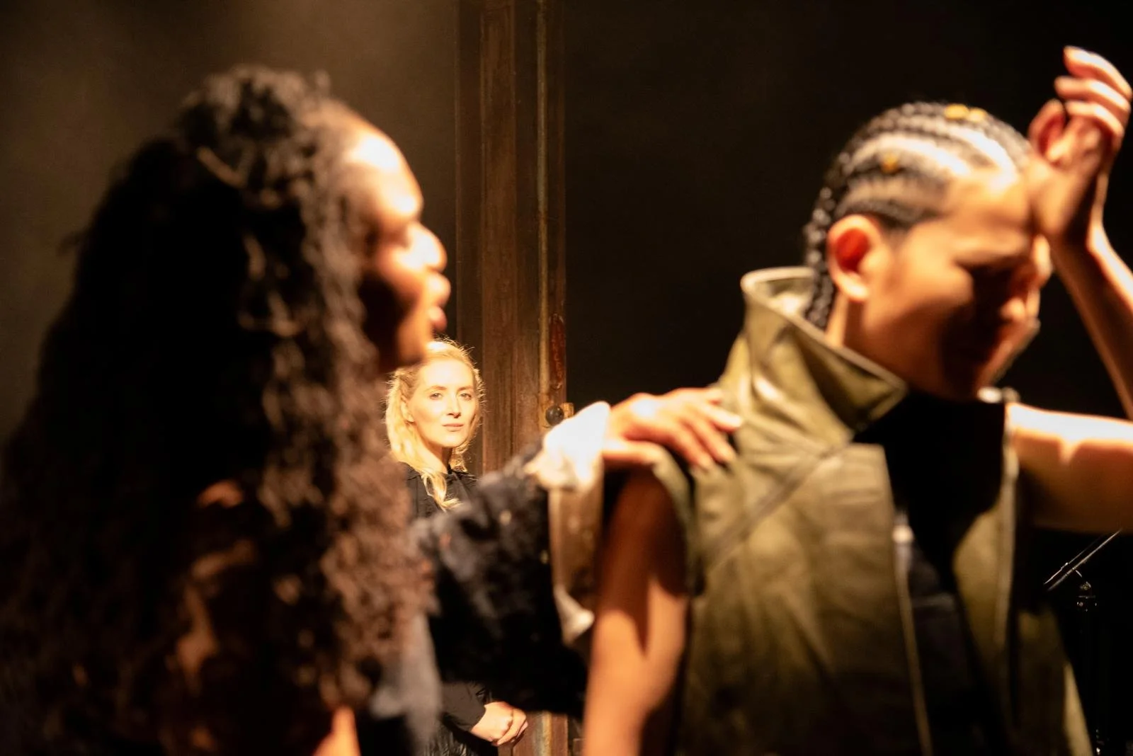 Two women are in the foreground, one with dark curly hair and the other with braided hair, both with their eyes closed and hands on each other's shoulders. In the background, a blonde woman is looking at them thoughtfully, standing near a wall.