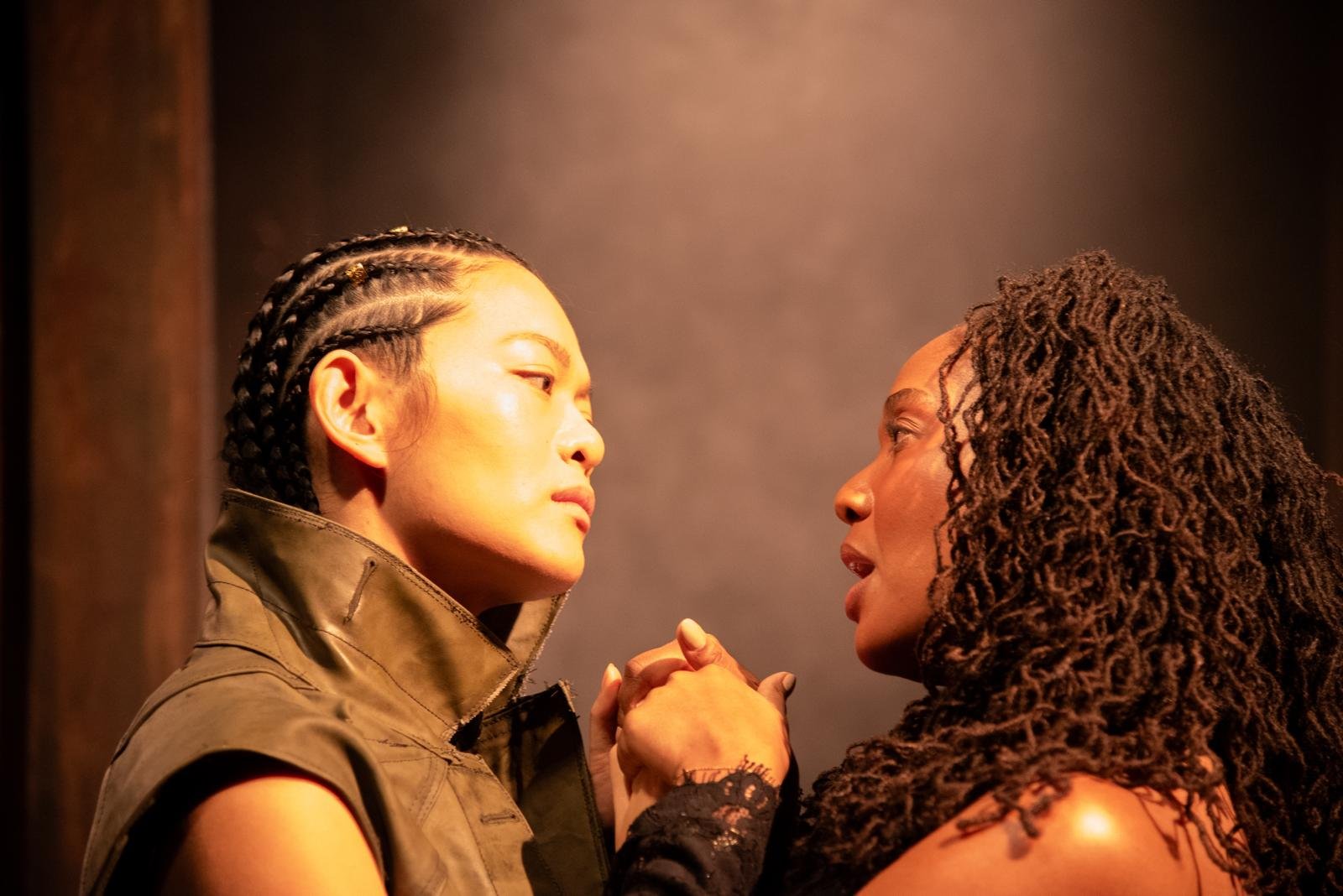 Two women face each other closely, holding hands, with intense expressions, in a dimly lit setting.