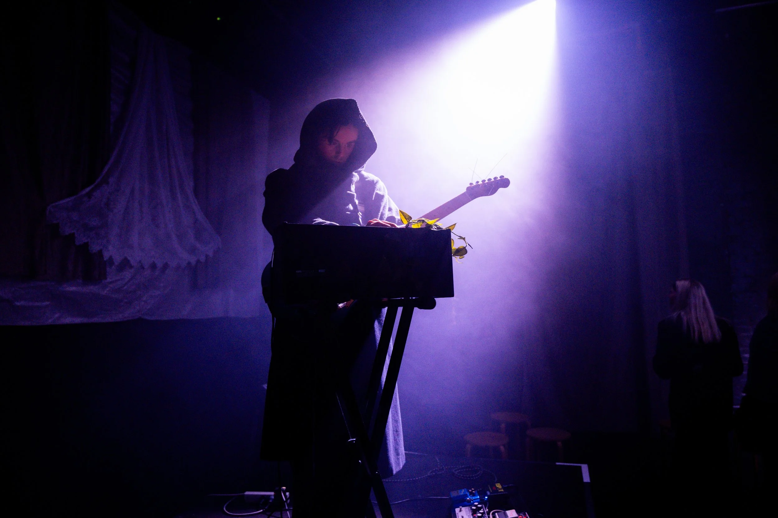 A person wearing a hoodie is on stage, playing an electric guitar and standing behind a keyboard, illuminated by purple and white stage lights, with a dark backdrop and some audience members in the background.