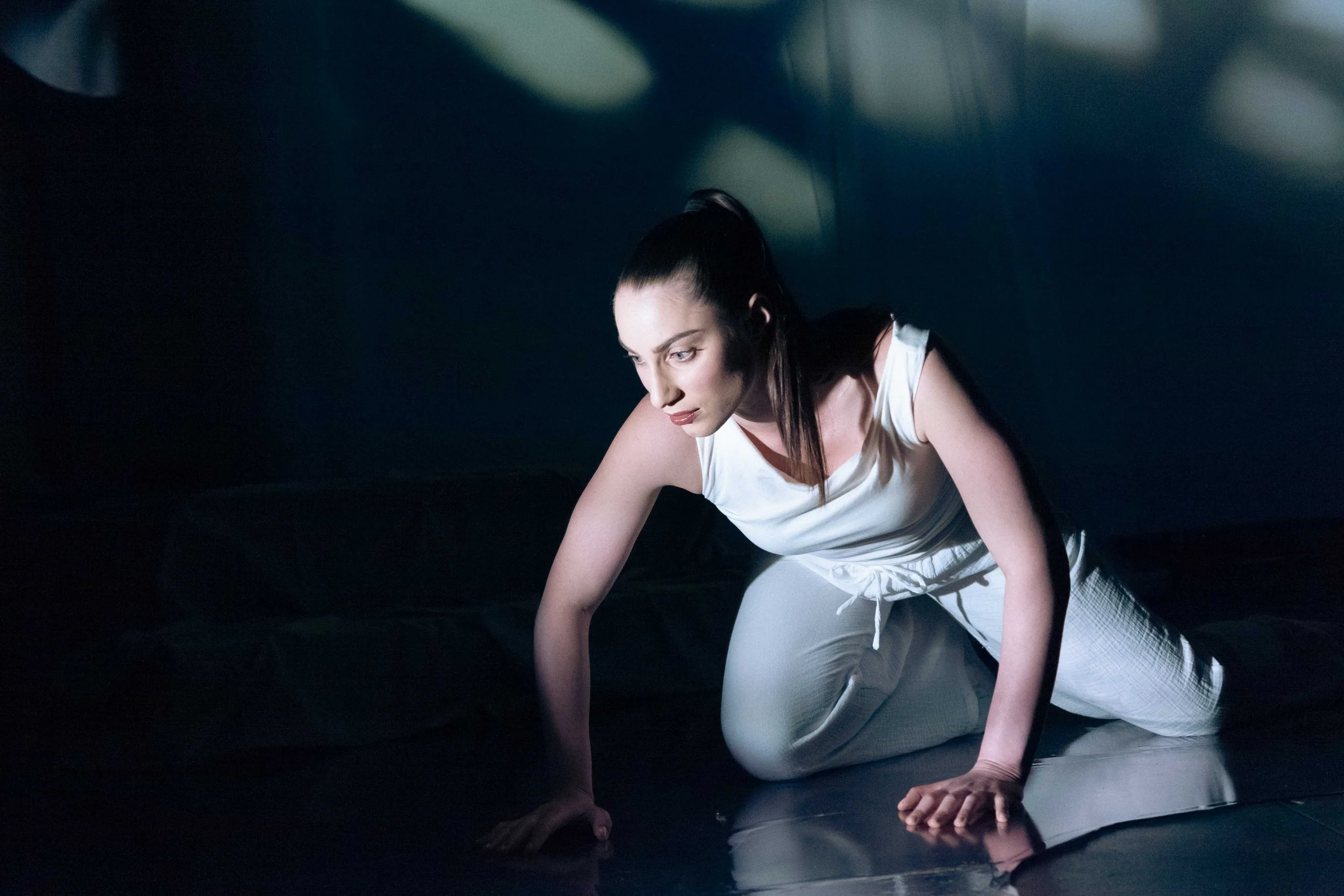 A woman with long dark hair tied back, wearing a white sleeveless top and light-colored pants, crouches on all fours on a dark, reflective surface in a dimly lit space, appearing focused.