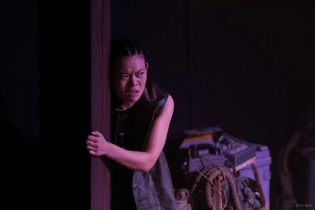 A woman peeks from behind a dark wooden door, looking concerned or wary, in a dimly lit room with various wires and equipment in the background.