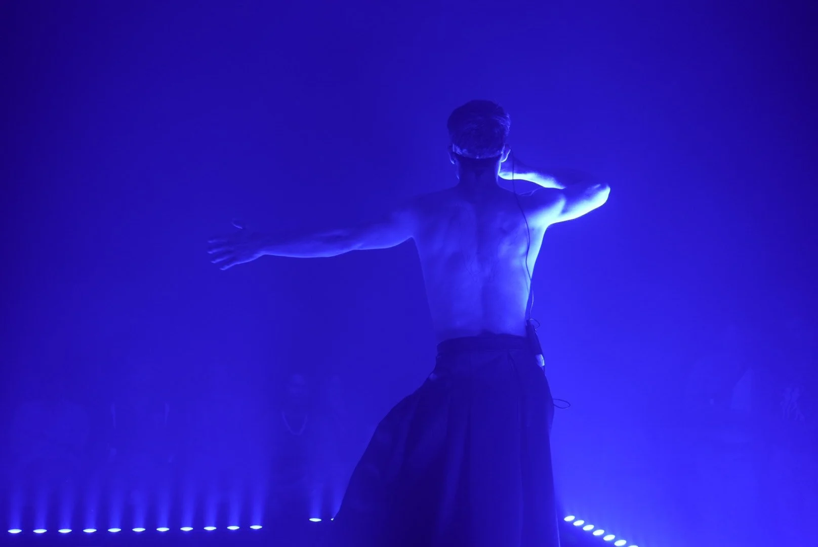 A shirtless man with headphones, with arms outstretched, standing in front of blue stage lighting, possibly performing or dancing.