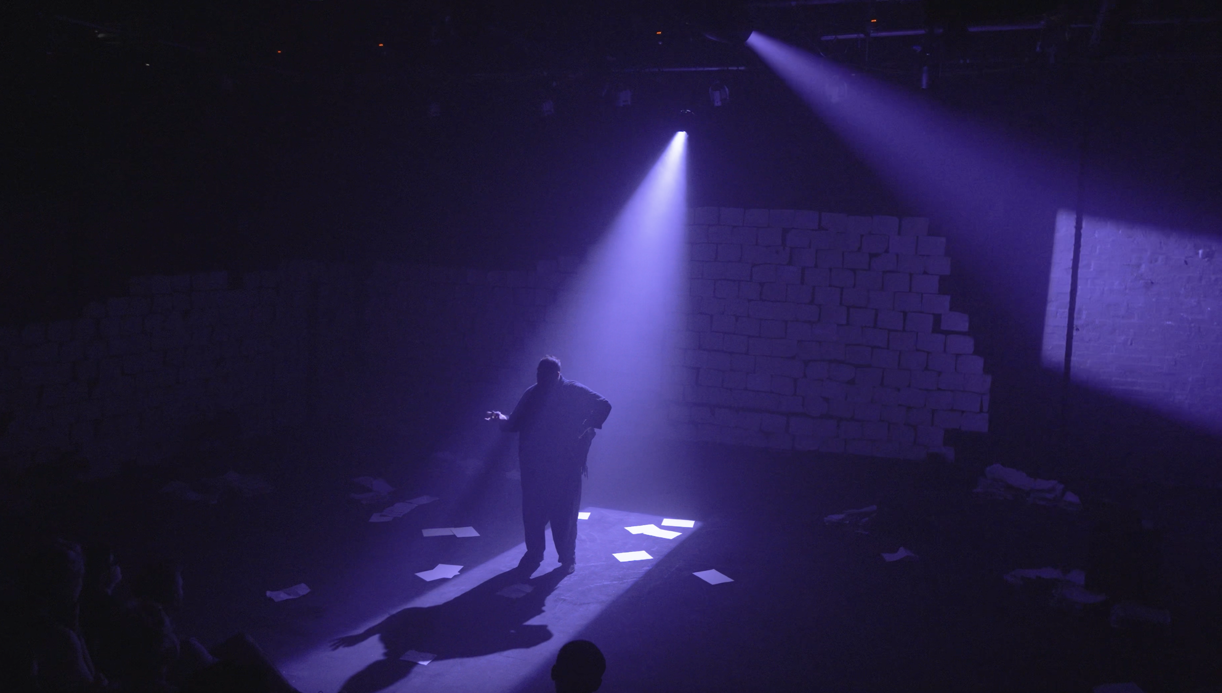 Stage with performer under purple spotlight, scattered papers on floor, brick wall backdrop, audience seated in darkness.