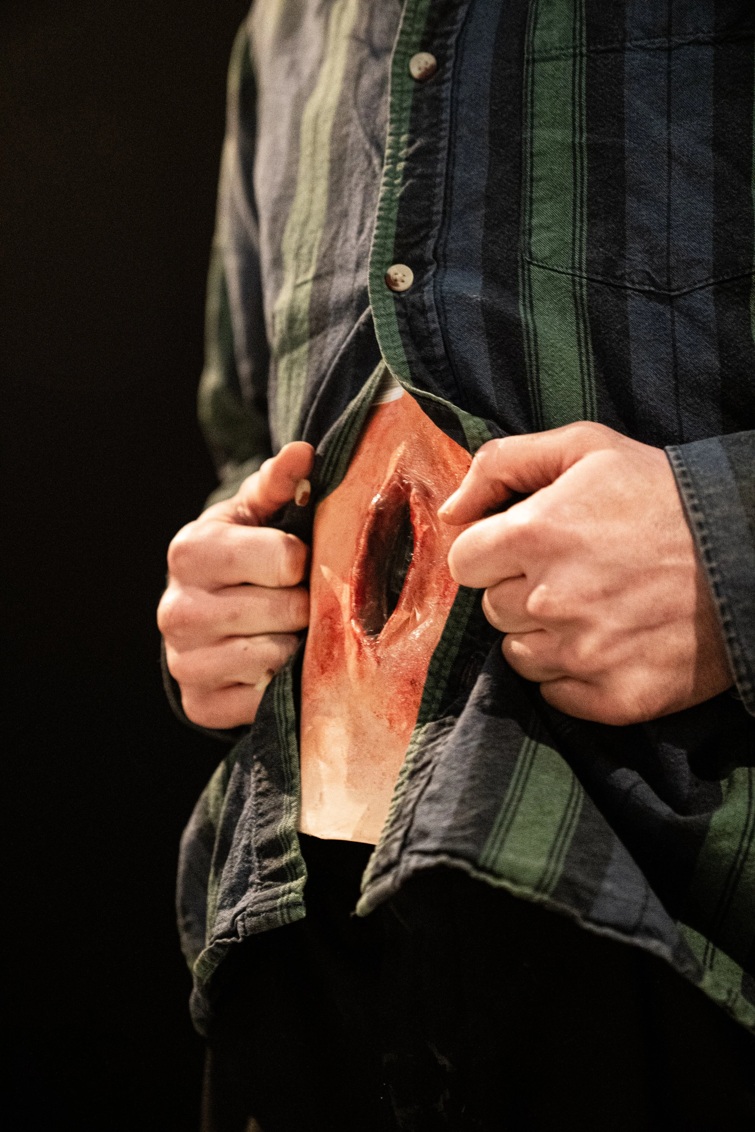 Person pulling up green and gray plaid shirt to reveal a large, realistic fake wound in the abdomen area.