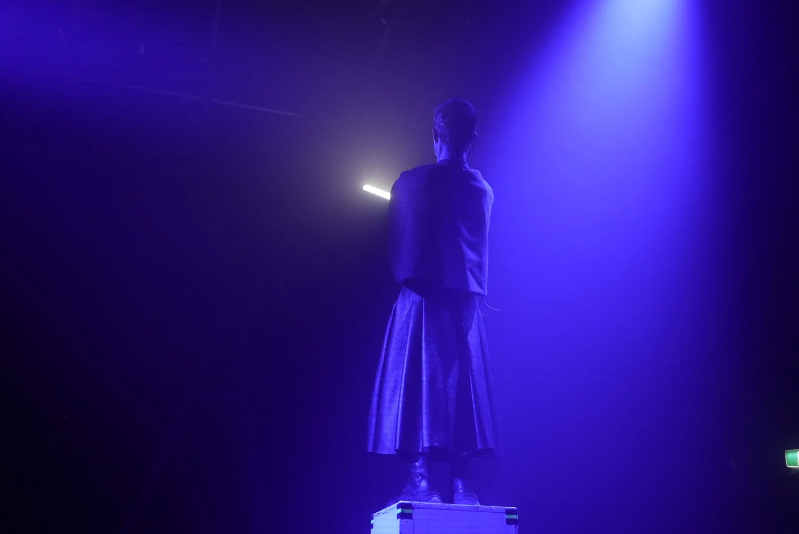 Person standing on a platform with arms crossed, illuminated by blue and purple stage lights, facing away from the camera.