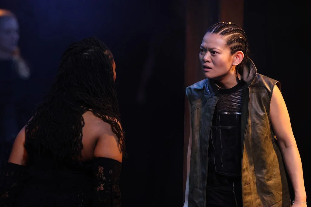 Two women having a heated conversation, one with braided hair and wearing a black leather vest, the other with long, loose dreadlocks, wearing a black dress.