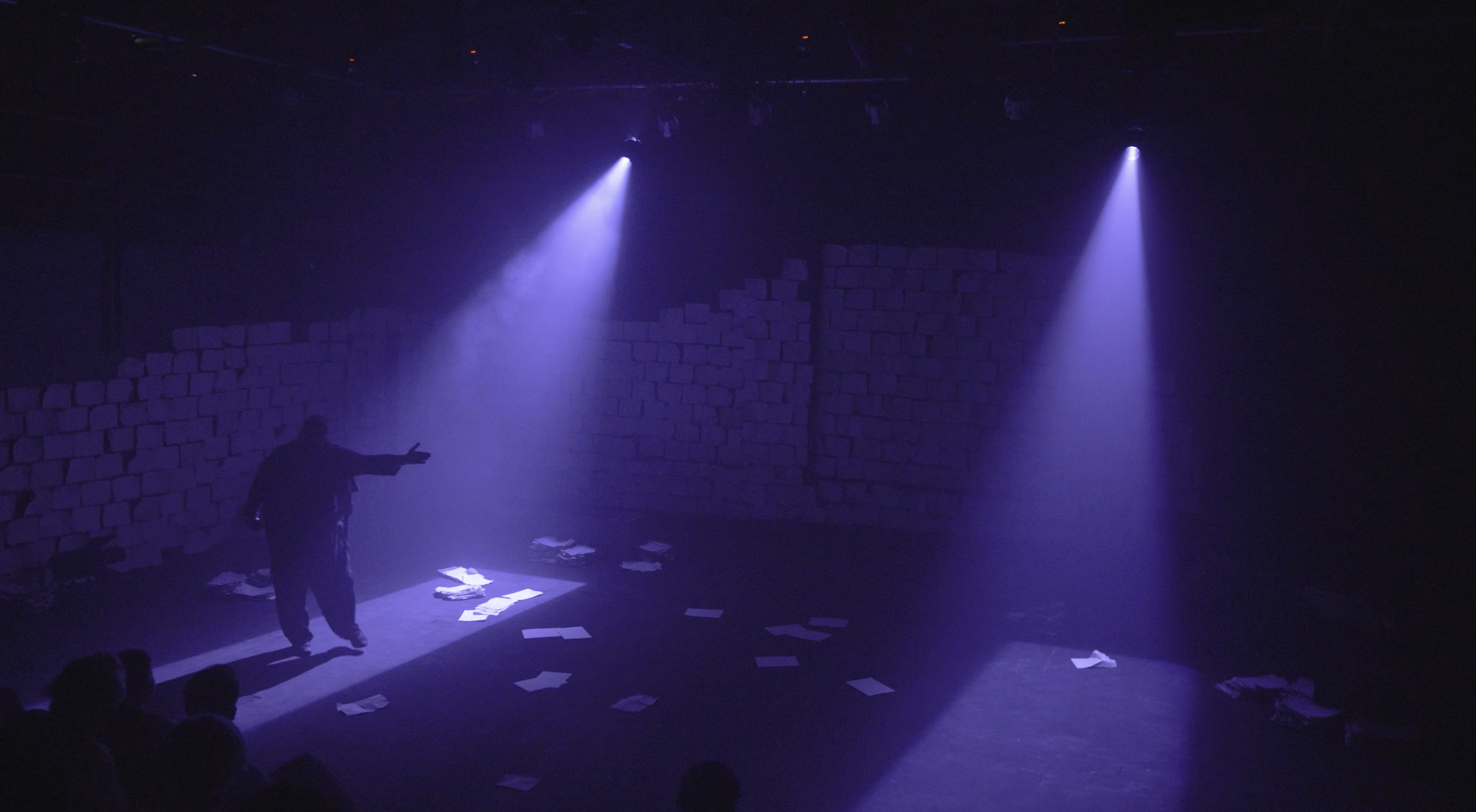 A stage with purple lighting, a person standing with outstretched arms, papers scattered on the floor, a brick wall backdrop, and an audience silhouette in the foreground.