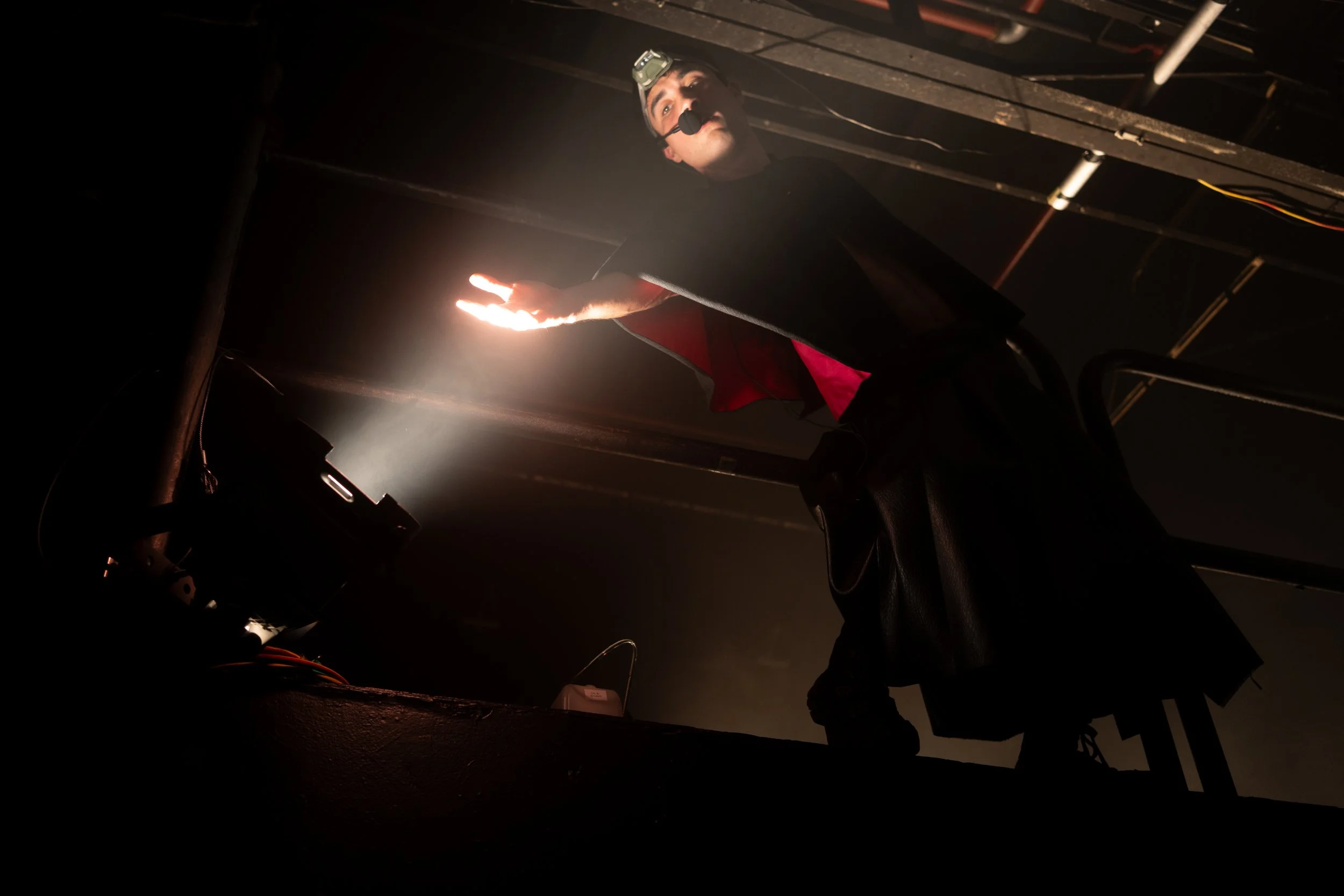 A person dressed as a magician performs a trick on stage, illuminated by a spotlight, wearing a headlamp and a costume with a black leather jacket and a red shirt, with a dark background.